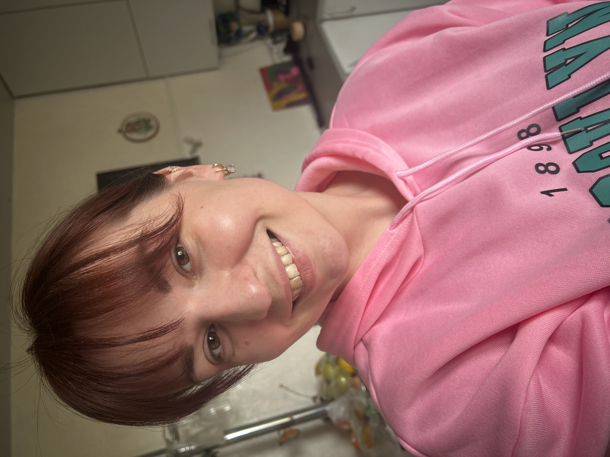 A medium shot captures a woman indoors, wearing a pink hooded sweatshirt with the text "1898" and another word in green lettering that is partially cropped out. Her hair is auburn with bangs, and she is smiling with a visible set of teeth. The background includes kitchen elements such as cabinets, a decorative plate on the wall, and various items on the counter. The environment suggests an indoor setting but lacks definitive indicators to pinpoint Фоки, Russia.