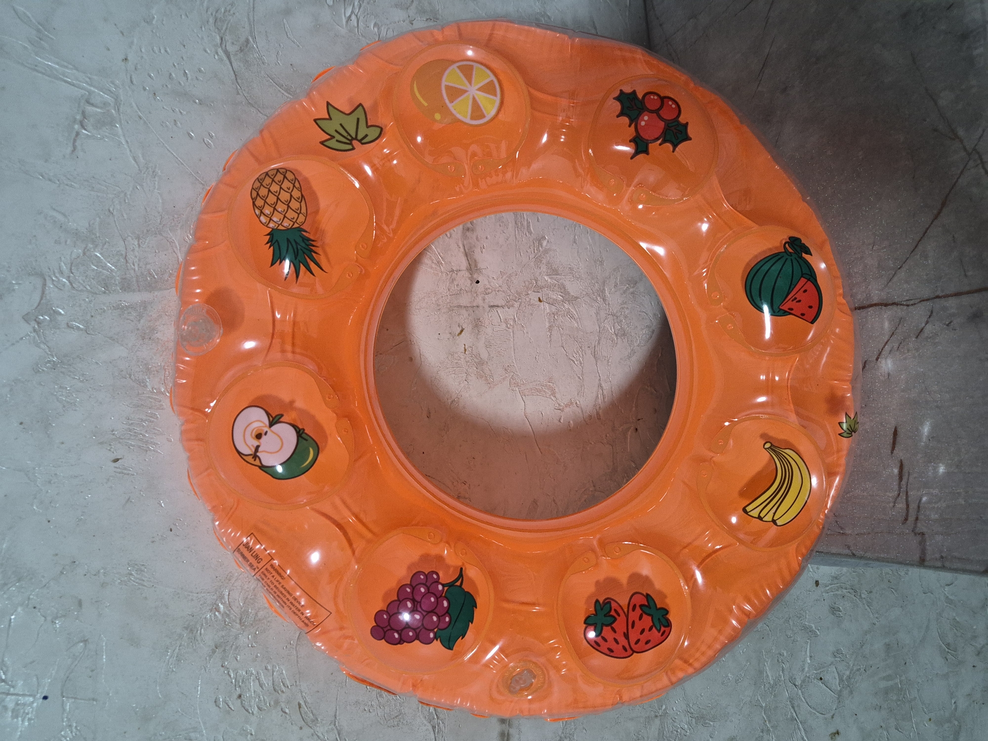 A close-up, overhead shot shows an inflatable orange swim ring with various fruit images printed on it. The ring is laid flat on a textured, grey surface. The fruits depicted include grapes, strawberries, bananas, watermelon, cherries, oranges, and a pineapple. The ring has a transparent quality, allowing the texture of the surface underneath to be partially visible. There are some small air valves visible on the surface of the ring. The background is a grey, textured wall. The location of this image in Қанабек, Kazakhstan, is not discernible from the visual information provided.