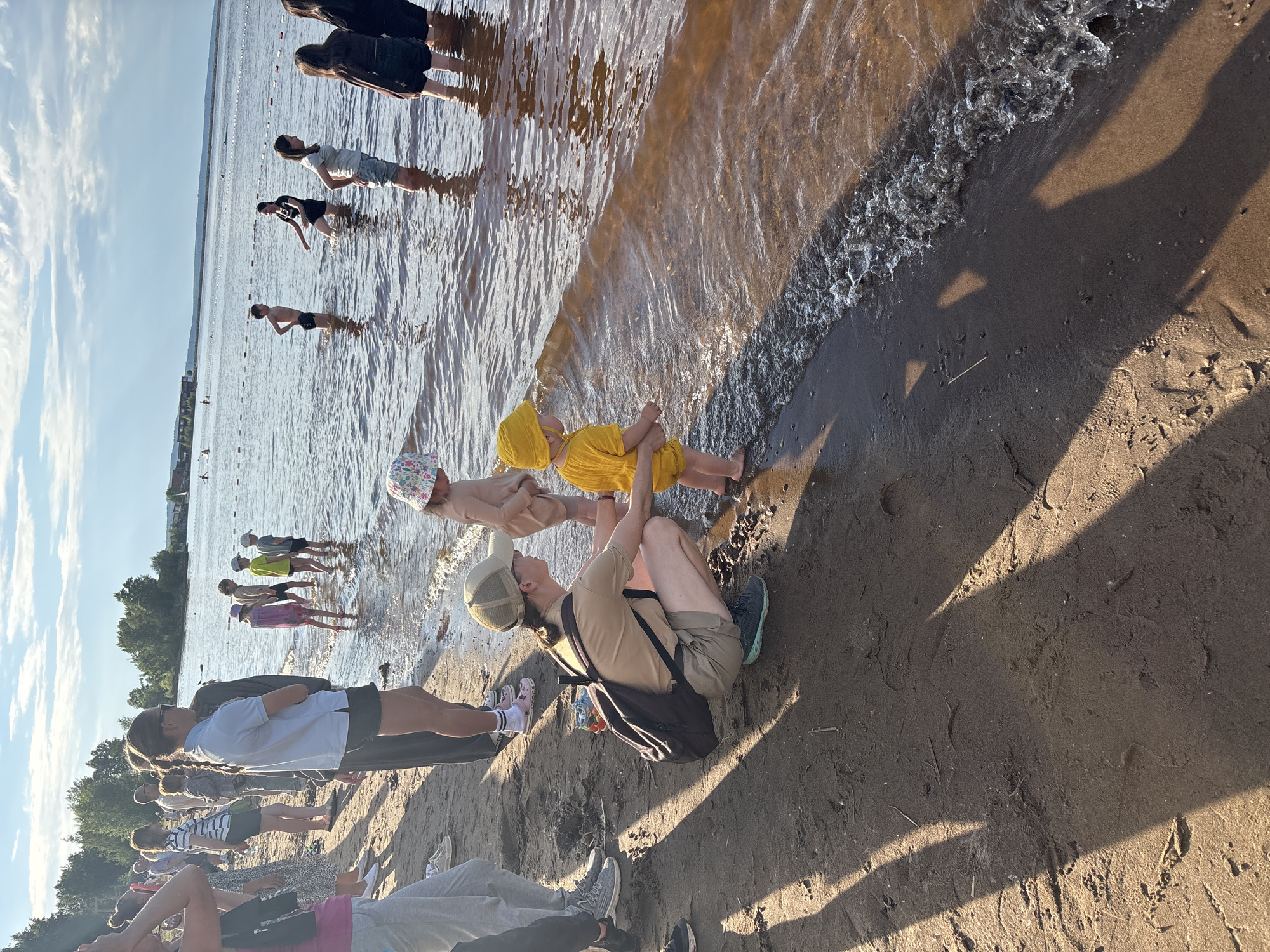 The image depicts a sunny day at a beach, likely located in Chaikovsky, Russia, near the Kama River. A group of people, including adults and children, are gathered on the sandy shore and in the shallow water. In the foreground, a woman wearing a tan cap and shorts is crouching down, facing a young child dressed in yellow. The child is standing at the water's edge, looking out at the river. Several other children and adults are visible further back, some wading in the water and others standing on the beach. The water is calm, with small waves lapping at the shore. The sky is blue with scattered clouds. The overall atmosphere appears relaxed and recreational.