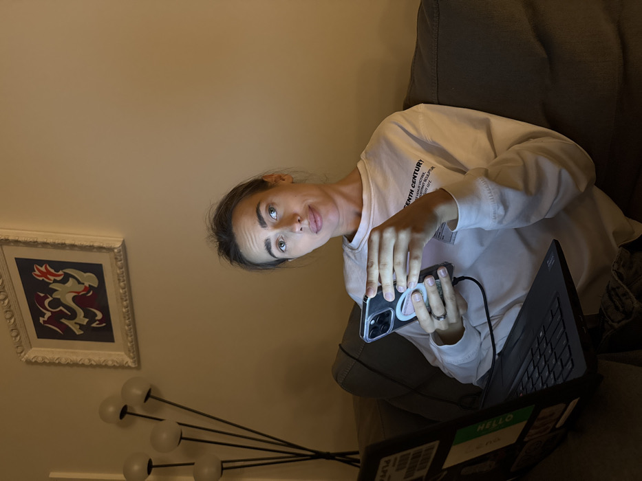 An indoor scene depicts a woman seated on a dark sofa, illuminated primarily by light sources external to the frame, creating a dim, warm ambiance. She wears a long-sleeved white sweatshirt, partially displaying text including "TENTH CENTURY" and "VENUS VICTRIX". A ring is visible on her left ring finger. She holds a smartphone with a clear case and a pink/white circular grip accessory, connected by a dark charging cable, manipulating it with both hands. Her gaze is directed upwards and to the right, with a slightly thoughtful or questioning facial expression. A dark laptop rests on her lap, open and emitting light from its screen, which shows a "HELLO" sticker on the left bezel. The background features a light beige wall. To the left, a modern floor lamp with five spherical, milky-white glass shades on black stems is visible. Above the lamp, a framed piece of abstract art hangs on the wall, featuring red, grey, and white shapes on a dark blue background within an ornate white frame. The interior setting provides no discernible visual cues to confirm the requested location in Venice, Italy.
