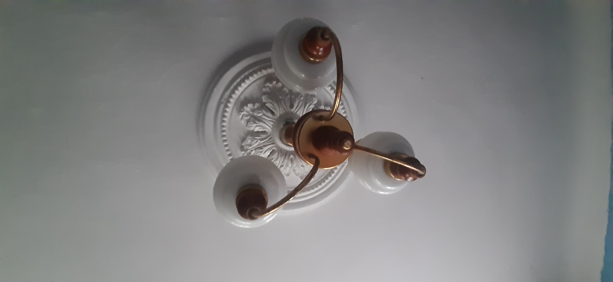 A ceiling-mounted, three-bulb light fixture with a decorative white rosette is photographed from below against a white ceiling. The fixture has a central brass-toned stem with wooden accents, and three curved arms extending outward, each holding a white glass bulb with a wooden decorative element at its base. The rosette appears to be made of plaster or a similar material. The image shows no people, actions, or interactions. The scene is indoors, likely in a residential setting, and there is no information to suggest it is located in Yessentukskaya, Russia.