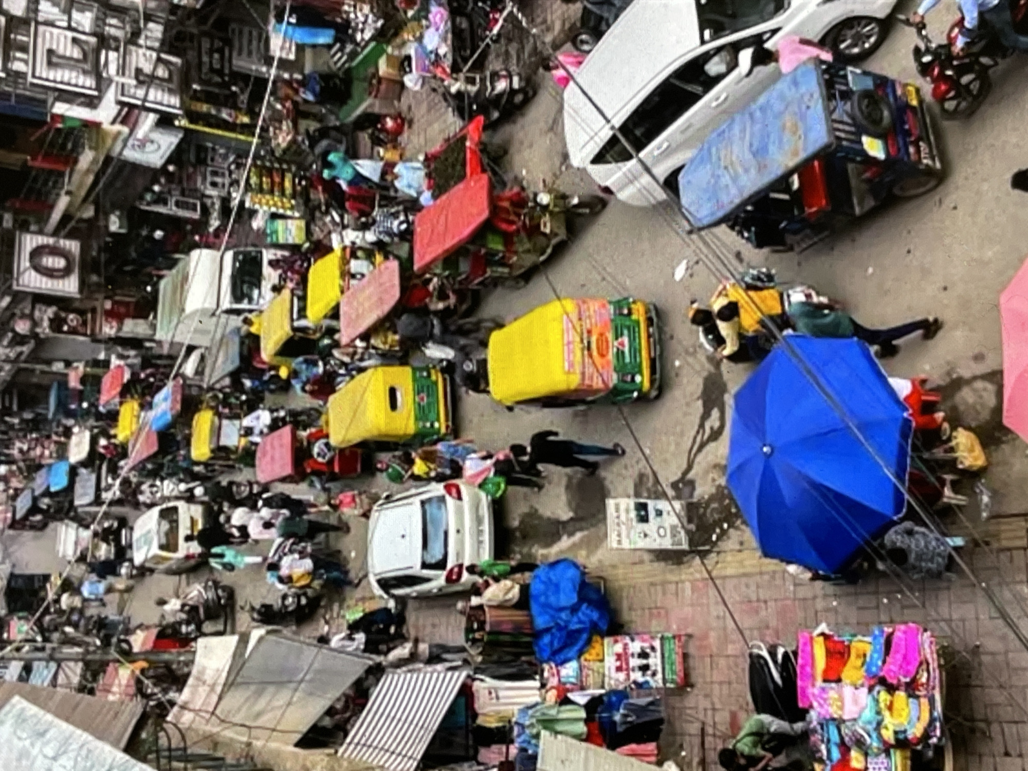 The image depicts a bustling street scene in a city, densely populated with vehicles and pedestrians. Multiple auto-rickshaws, some yellow and green, others red and yellow, are visible, indicating they are a primary mode of transport. Several motorcycles and a white car are also present. Pedestrians are walking along the narrow street, navigating between the vehicles and market stalls. The stalls appear to be selling various goods, including clothing and other items, with some displaying colorful fabrics. A large blue umbrella is open, suggesting a temporary shelter or vendor. The overall environment appears to be an urban market or commercial area, characterized by a high level of activity and congestion. The question of the location being in the city of Yalga, Russia, cannot be answered based on the visual information present in the image, as there are no identifying markers, signs, or architectural features that would specify this location.