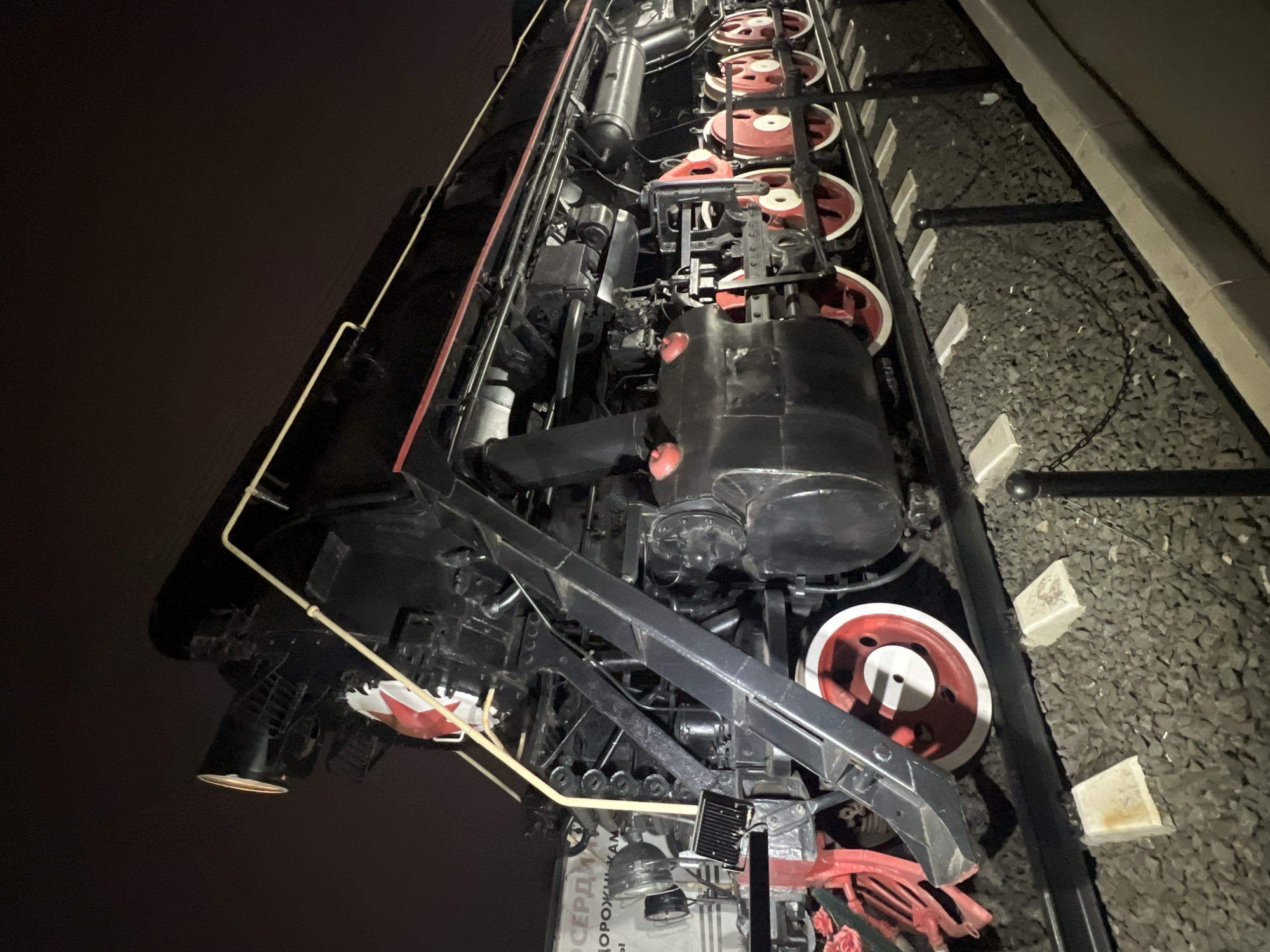 The image shows a close-up, angled view of a black and red steam locomotive, located on a section of railroad track laid on gravel. The scene appears to be at night or in low light. The locomotive has numerous wheels visible, some with red painted centers. A portion of a building or structure is visible at the right side of the image, with a concrete or stone edge. The photograph seems to be taken at a low angle, looking up toward the machine. A plaque with Russian lettering can be seen attached to the front of the locomotive. Based on the inscription, the locomotive is located in Cherepovets, Russia.
