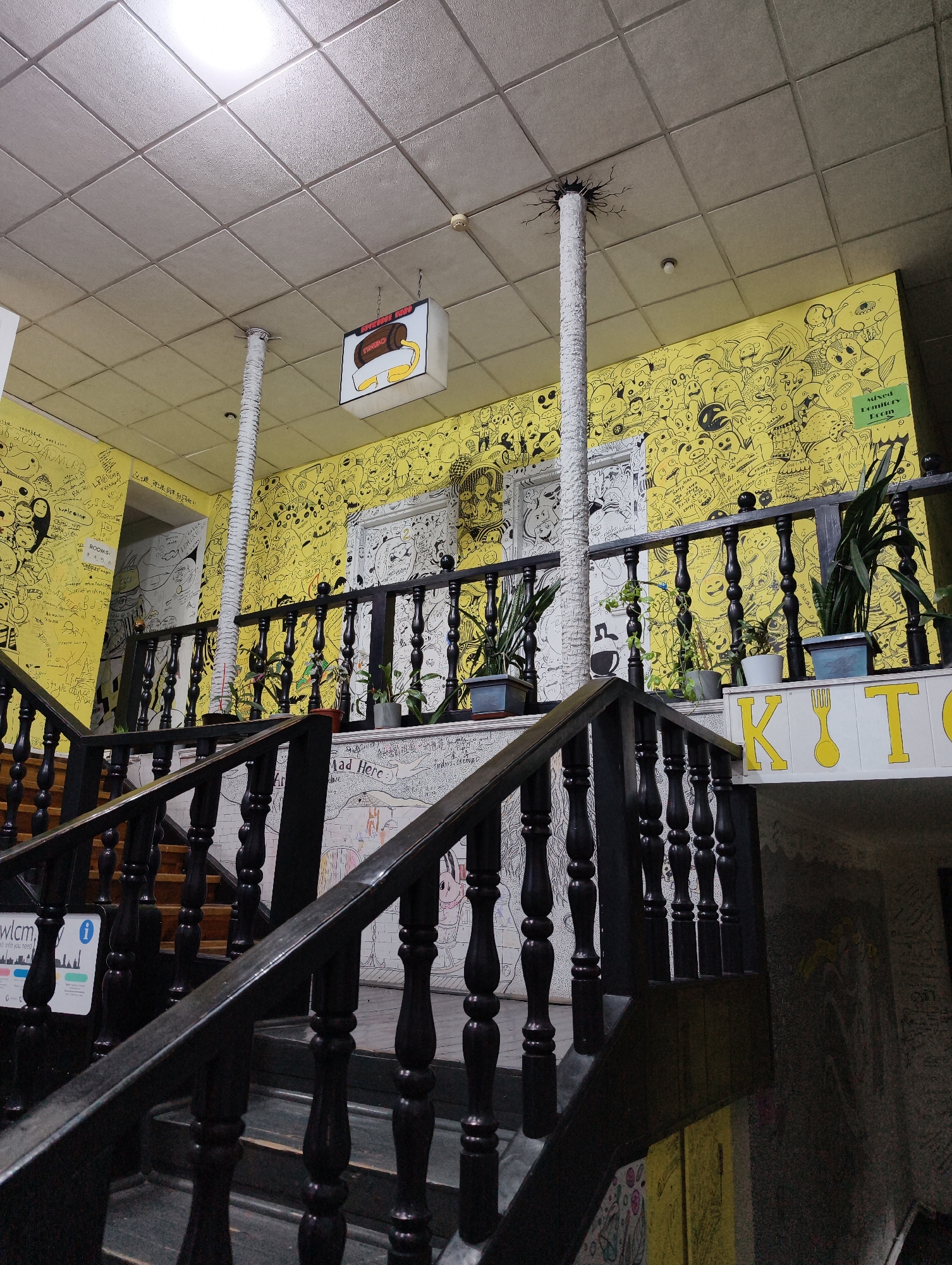The image shows the interior of a building with a staircase. The walls are decorated with black and white drawings on a yellow background. A black wooden railing runs along the staircase. Several potted plants are placed on the railing. A sign saying "KITO" is visible in the background. A light fixture is suspended from the ceiling, and a decorative column extends to the ceiling. Signs displaying information are present, including one with "wlcm" and other symbols, and one with "Mad Here." Text reading "2024ADMIRE," "ROOMS:" and "Welcome" are visible on the walls. The image captures a creative and unique interior design, suggestive of a public space in Tashkent, Uzbekistan.