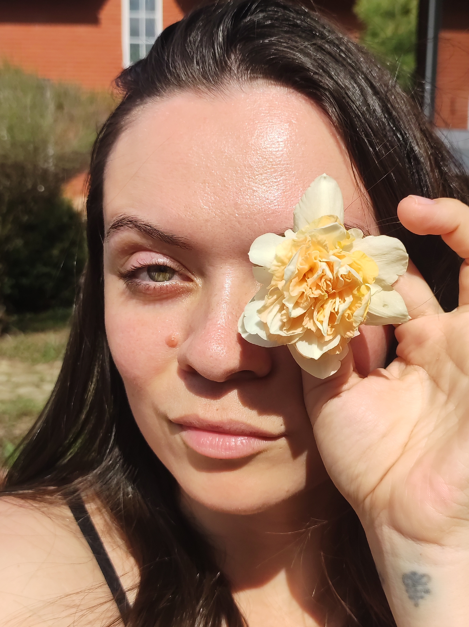 A woman with dark brown hair is holding a yellow and white flower in front of her right eye. She has fair skin with a mole near her nose. Her left eye is visible and appears to be hazel or green. Her lips are light pink. She is wearing a black strap, suggesting she is wearing a tank top or similar garment. A faded tattoo is visible on her left wrist. The background shows a brick building with a white-framed window and some greenery. The setting appears to be outdoors during the daytime, likely in sunny weather. The location is Фоки, Russia.