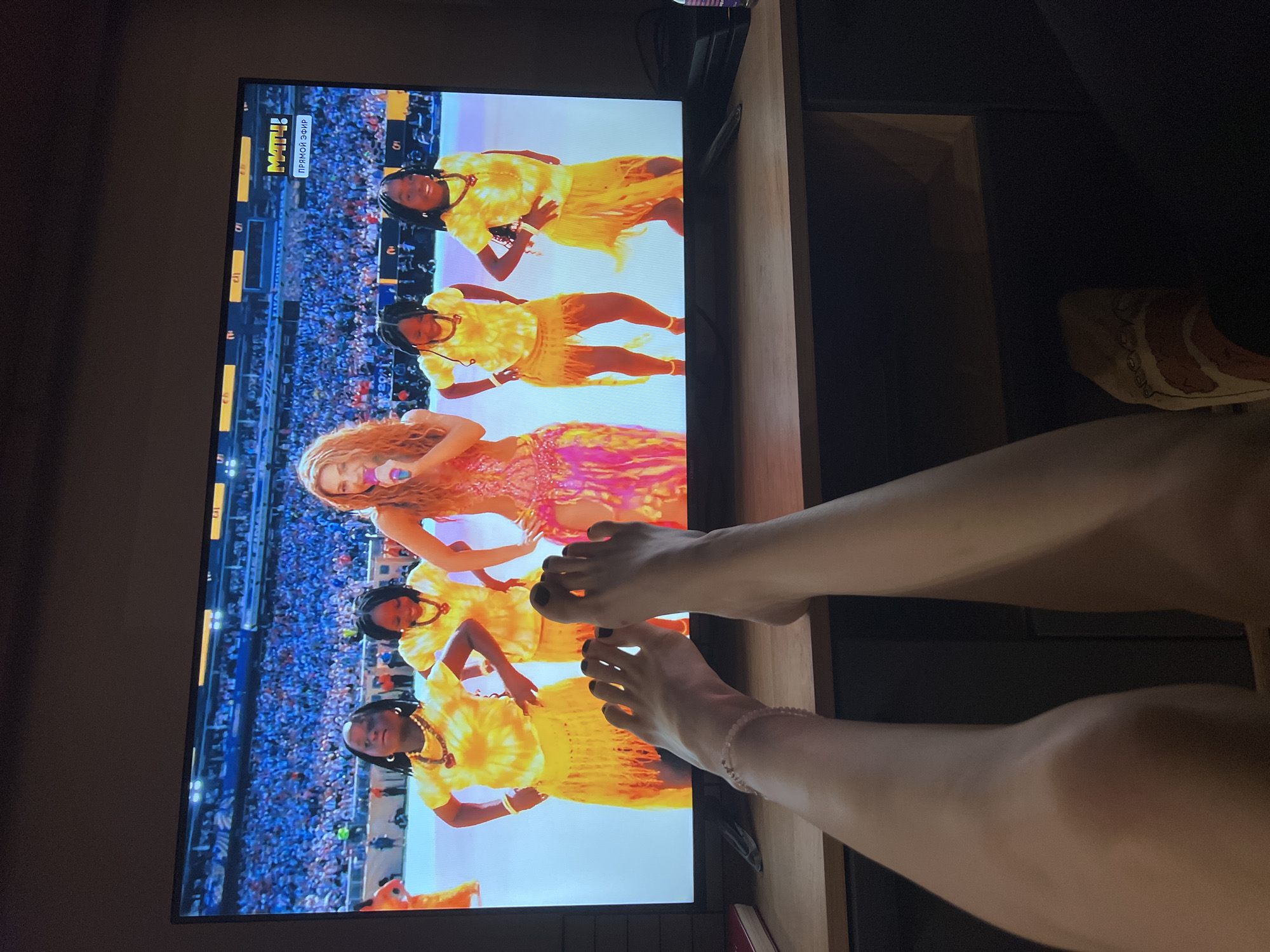 A wide-screen television displays a live performance on a stage in front of a large, cheering crowd. A female performer, with long, wavy blonde hair, wears a sparkling, form-fitting pink and gold dress and holds a microphone. She is accompanied by four young girls dressed in matching yellow tie-dye tops and yellow fringed skirts, who are dancing. A "МАТЧ!" logo with "ПРЯМОЙ ЭФИР" (Live Broadcast) text is visible in the upper right corner of the screen, suggesting the broadcast is from the Match TV channel. In the foreground, a person's bare legs and feet are visible, positioned in front of the television. The feet are adorned with black nail polish and one foot wears a delicate beaded anklet. The television is placed on a wooden desk, with a few objects partially visible on the left side, including a book and a pen. The background is dimly lit, indicating the room is dark. The scene appears to be a living room or entertainment space, likely in Moscow, Russia, given the presence of the "МАТЧ!" broadcast logo, which is a prominent Russian sports television channel.