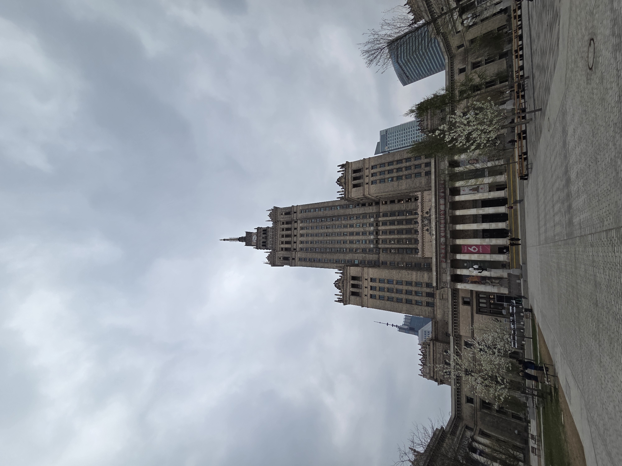 A low-angle, outdoor shot shows the Palace of Culture and Science in Warsaw, Poland, on a cloudy day. The building, a tall Stalinist-style skyscraper, dominates the frame, extending upwards towards the overcast sky. The architecture features multiple setbacks and spires. A paved walkway extends from the right side of the image, with trees and smaller buildings visible along the base of the Palace. Some people are walking near the building. A modern, glass skyscraper is partially visible behind and to the right of the Palace.