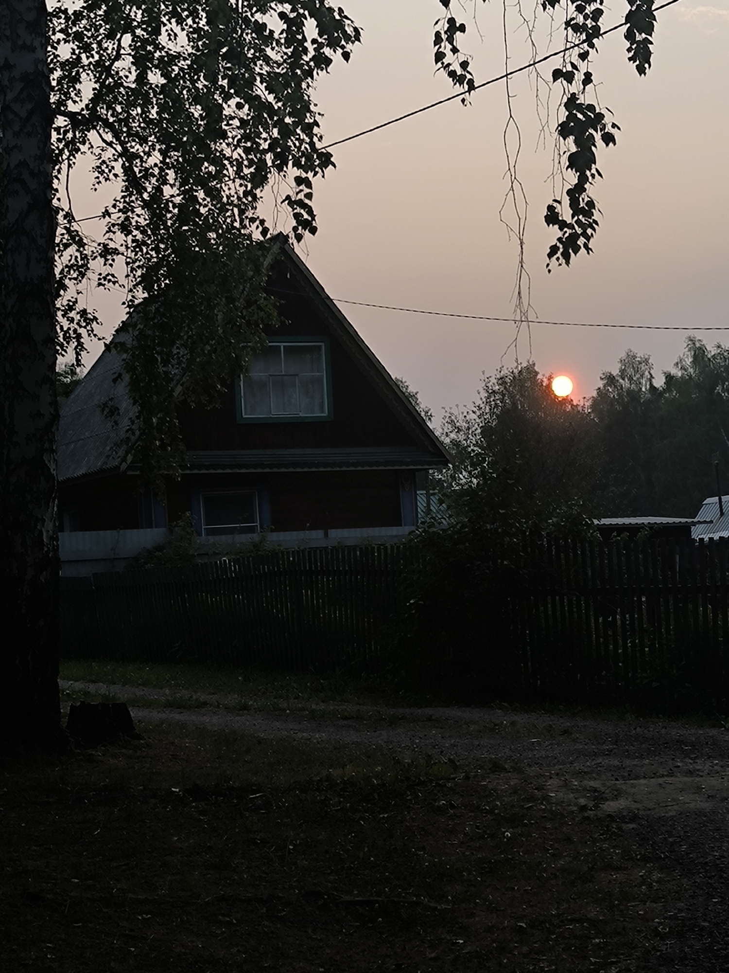This media file captures a rural scene in Maiskii, Russia, during sunset. The primary subjects are a traditional wooden house with a steep roof and a weathered wooden fence. The house appears to be a dacha, a common type of summer house or cottage in Russia. Hanging branches with leaves, likely from a birch tree given the bark visible on the left, frame the upper portion of the image. In the background, a dense line of trees forms a dark silhouette against the hazy, orange-tinged sky.
The sun is a vibrant, glowing orb low on the horizon, partially obscured by the trees, indicating the time of day is sunset. The sky suggests a warm, possibly humid atmosphere, with the haze potentially caused by dust, smoke, or humidity. The overall mood is serene and peaceful, evoking a sense of quiet twilight in the countryside. The foreground shows a dark, unpaved path and grassy areas, with a tree stump visible near the large tree trunk on the left. The scene is devoid of any people or overt activity, emphasizing the stillness of the moment. No visible text is present in the image.