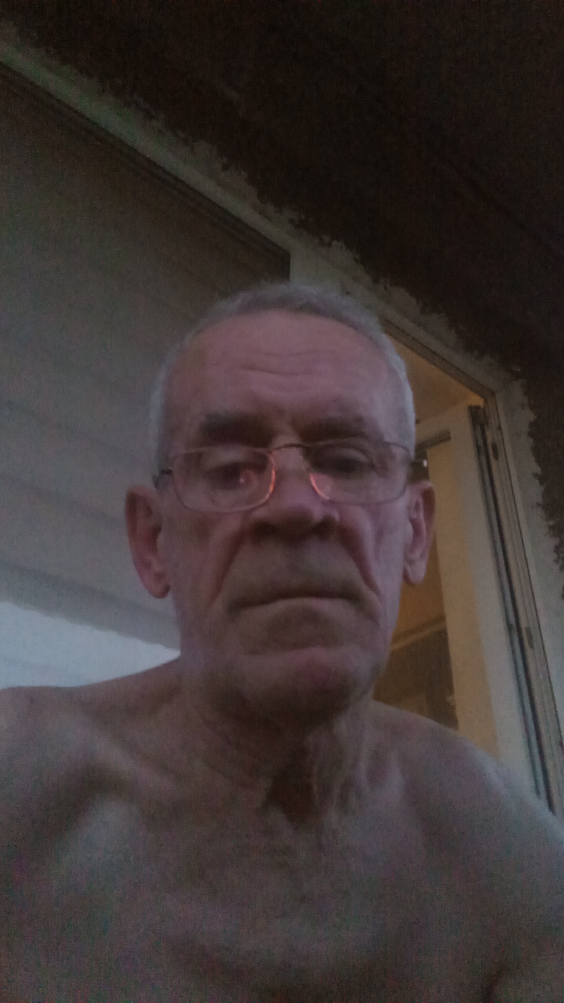 A close-up, waist-up, selfie-style photograph depicts an elderly Caucasian male with graying hair and fair skin. He is wearing glasses with thin wire frames. The man's face is turned slightly to the left, and his mouth is closed in a neutral expression. His bare shoulders and upper chest are visible, suggesting he is not wearing a shirt.
The background is dimly lit and appears to be an outdoor setting, possibly a porch or patio. To the left, a light-colored, possibly wooden, wall or siding is visible, and to the right, a doorway or window frame can be seen, with a glimpse of a light-colored interior beyond. The overall lighting suggests it might be dusk or early morning.
There is no identifiable information within the image to suggest a specific location within the city of Tiachiv, Ukraine.