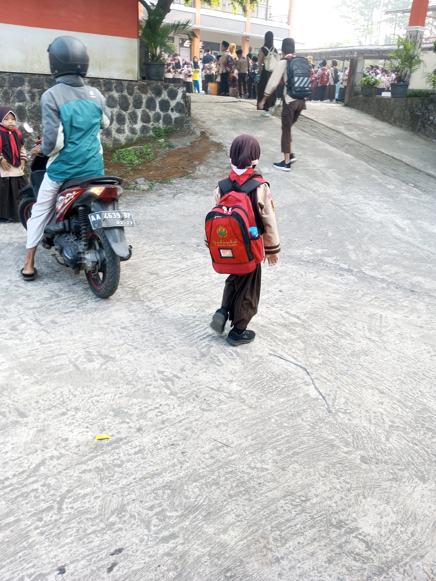 In Krasak, Indonesia, a young student wearing a red backpack and brown uniform walks away from the camera on a concrete path. The student is facing right and is captured from behind. In the background, a person on a red motorcycle, wearing a helmet and a gray and teal jacket, is stationary. Behind the motorcycle rider, a smaller child in a brown uniform and red headscarf is visible. The environment appears to be an outdoor area, possibly a school campus, with a building and trees visible in the distance. The concrete path has a textured, weathered surface.