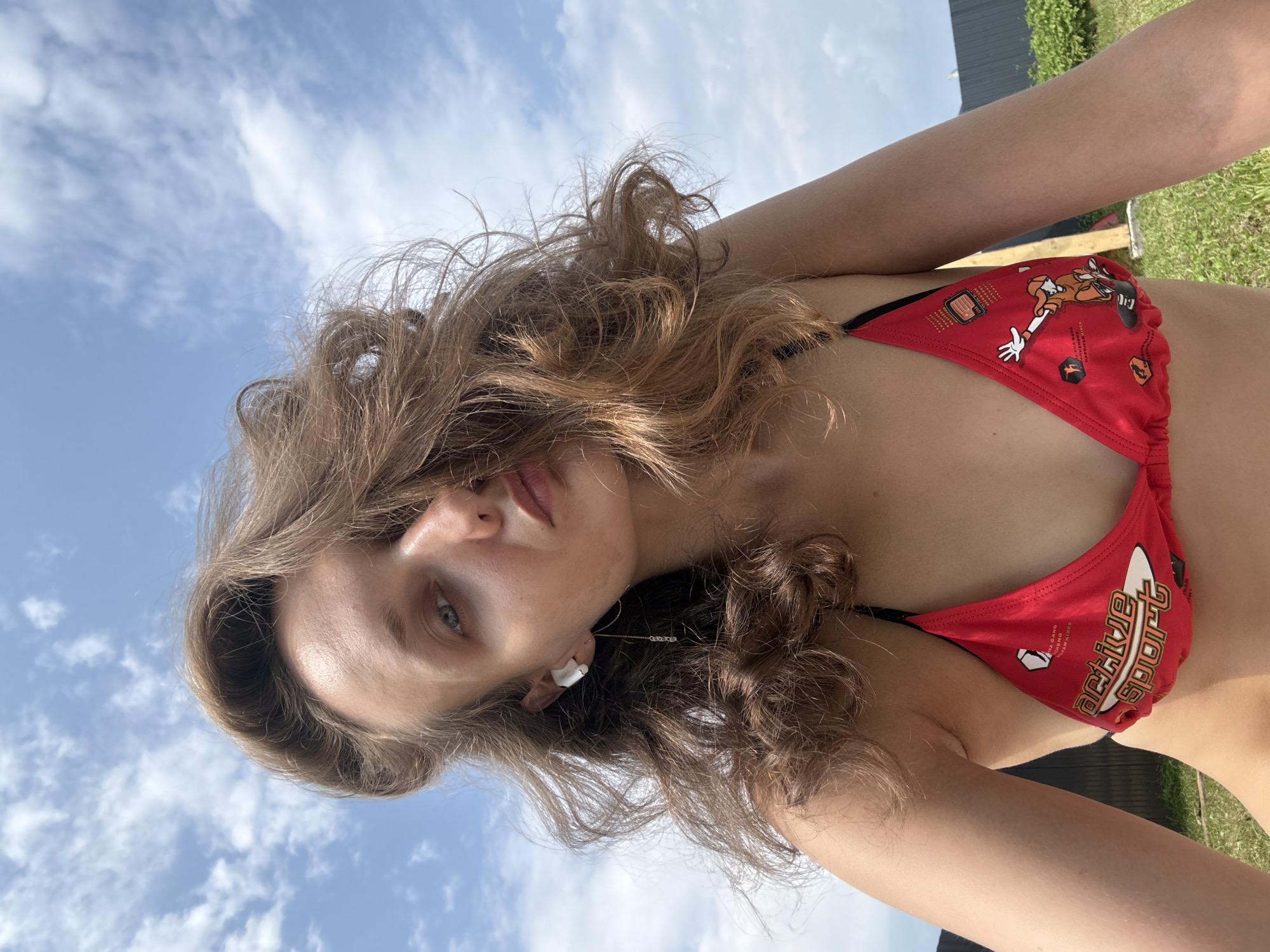The image is a selfie of a young woman outdoors on a sunny day. She is wearing a red bikini top with a cartoon design. Her long, wavy brown hair is falling over her face and shoulders. She has a white earbud in her left ear with a dangling chain earring. The background shows a blue sky with white clouds, a wooden fence, and some green grass and foliage. The woman is looking directly at the camera with her mouth slightly open. The image appears to be taken in the village of Podshivalovo, in Russia.