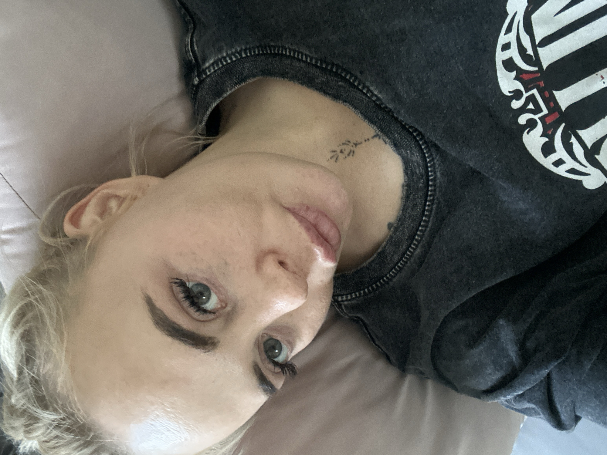 A fair-skinned individual with light blonde hair lies on a light-colored surface, likely a pillow or bedding. The person is wearing a dark grey crewneck sweatshirt with a design on the right shoulder. A small, dark tattoo is visible on the lower neck. The individual has green eyes and is looking toward the camera. The location in the city of Vysoky, Russia is not discernible from the image.