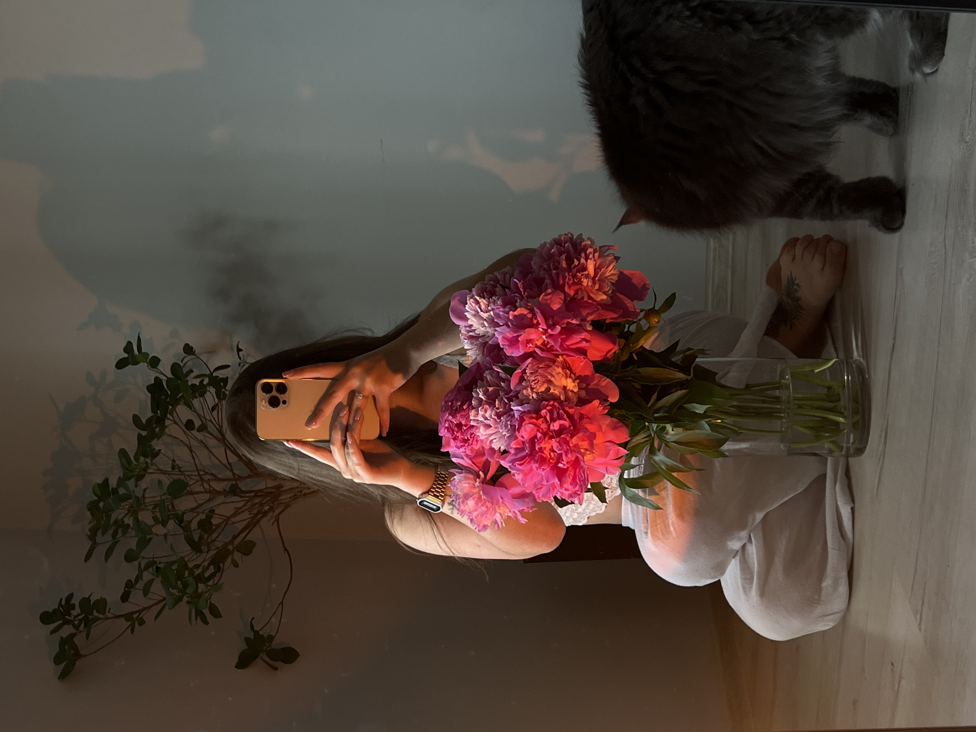 A young woman is captured in a selfie with a bouquet of pink peonies. She is kneeling on the floor, holding a gold smartphone in her hands, and her face is obscured by the device. She is wearing a white bralette and white pants, with a gold watch on her left wrist. The peonies are arranged in a clear glass vase, which sits on the floor beside her. To the right of the woman, a fluffy grey cat is visible, with its back to the camera. The background features a potted plant with green leaves casting shadows on a light gray wall. The scene is illuminated by warm, directional light, creating a soft ambiance. There is no discernible location in Ternopil, Ukraine, mentioned or implied within the image.