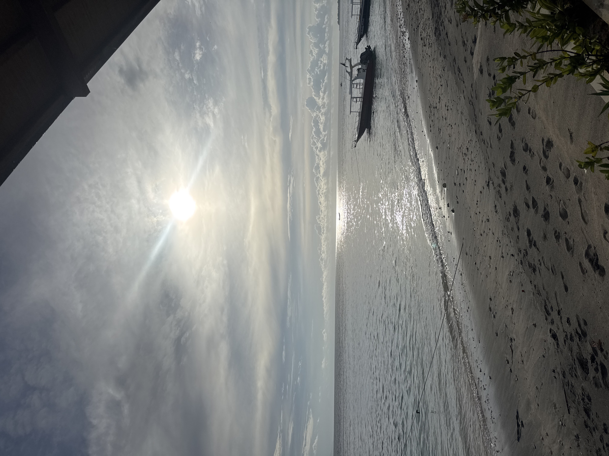 The image shows a beach scene in Nusa Penida, Indonesia. The sun shines brightly in the sky, partially obscured by clouds. To the right, a sandy beach with footprints leads to the water's edge. Small waves lap at the shore. A boat is anchored in the water near the beach. Branches of a green plant are visible in the upper right corner, and a building element is at the upper left.