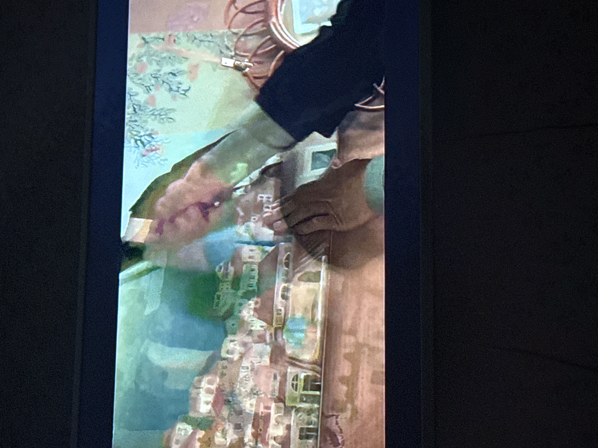 A close-up photograph shows a hand placing what appears to be a model building onto a painted, topographical map or landscape. The building seems to be one of many forming a miniature settlement. The arm and partial view of a person's attire (dark sleeve) are visible, suggesting a person interacting with the scene. The overall image appears to be a photograph of a display or screen rather than the physical environment being represented.