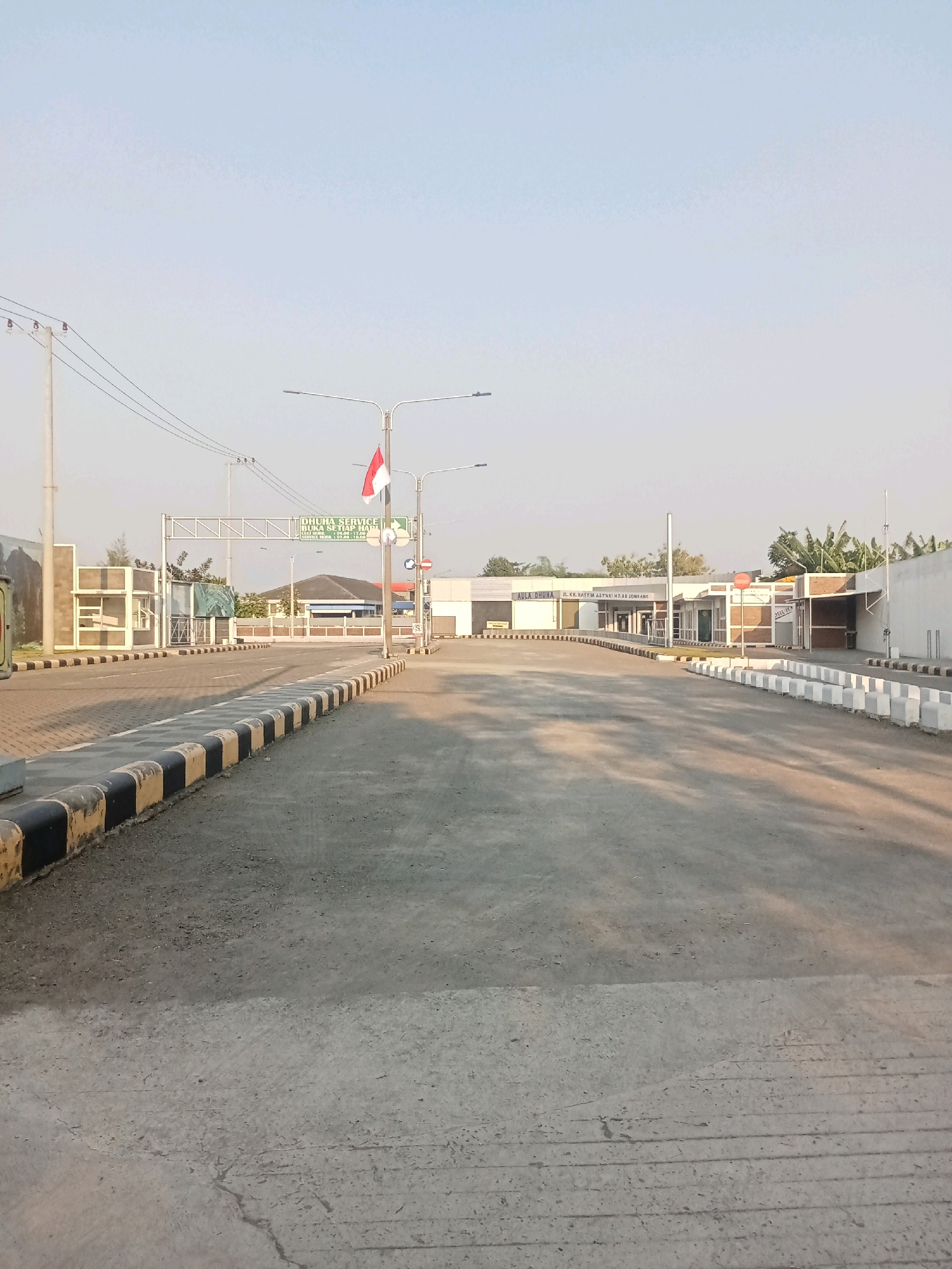 The image depicts an outdoor scene at what appears to be an entrance or checkpoint in Kaliwungu, Indonesia. The foreground is dominated by a paved road with distinct black and yellow striped curbs lining its edges. In the midground, there is a wide paved area leading towards a cluster of buildings.
One prominent building has signage that reads "AULA DHUNA JL. KH. HASYIM ASY'ARI NO. 88 JOMBANG". Another sign above the road indicates "DHUNA SERVICE BUKA SETIAP HARI". The buildings are low-rise and appear to be constructed with white walls and some exposed brick elements. There are several streetlights visible throughout the scene.
A red and white flag, likely the Indonesian flag, is flying on a flagpole to the right of the entrance signage. The environment suggests a sunny day with a clear sky. The overall impression is of an organized facility, possibly a service center or administrative complex, situated along a main thoroughfare. No people are visible in the image.