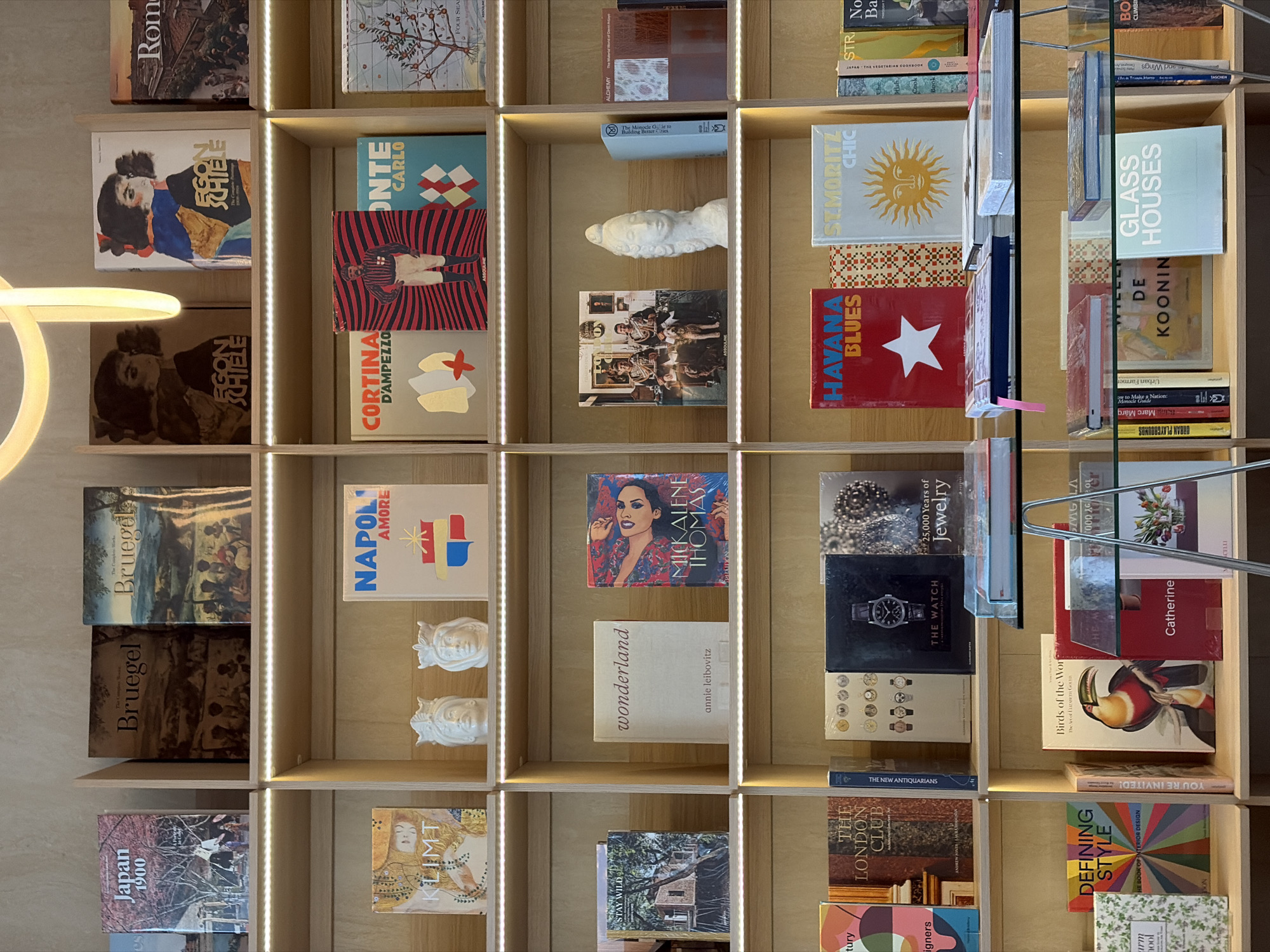 This image shows a wall of bookshelves. The shelves are filled with books of various sizes and colors. Some of the books are stacked horizontally, while others are standing upright. The books on the shelves include "Japan 1900", "KLIMT", "Bruegel", "Egon Schiele", "Napoli Amore", "Cortina D'Ampezzo", "Mickalene Thomas", "Wonderland" by Annie Leibovitz, "The London Club", "Defining Style", "The New Antiquarians", "You're Invited!", "25,000 Years of Jewelry", "The Watch", "Birds of the World", "Catherine", "Monte Carlo", "Havana Blues", "St. Moritz Chic", "Glass Houses", "De Kooning", and "Urban Playgrounds". There are also some decorative objects on the shelves, including two small white sculptures of human faces. The shelves are illuminated by a soft light, which highlights the colors and textures of the books. The location appears to be a bookstore or library.