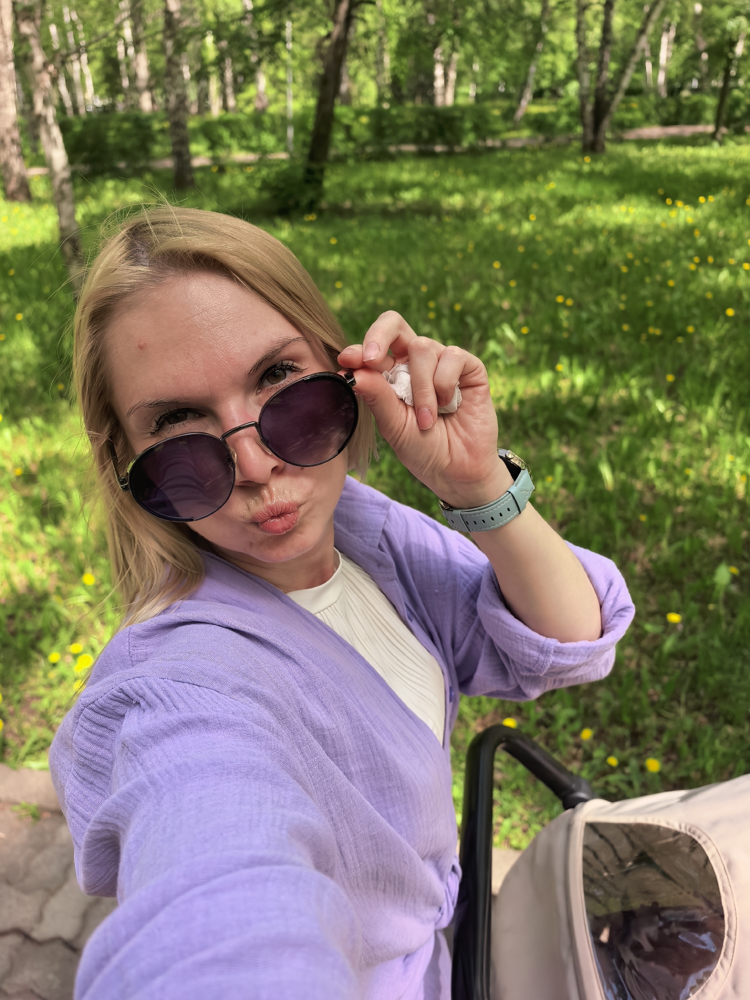 A woman is taking a selfie in a park in Sterlitamak, Russia. She is holding her sunglasses and making a kissing face. She is wearing a lavender shirt and a light blue watch. In the background, there is a green lawn with dandelions and birch trees. A stroller is visible in the bottom right corner.