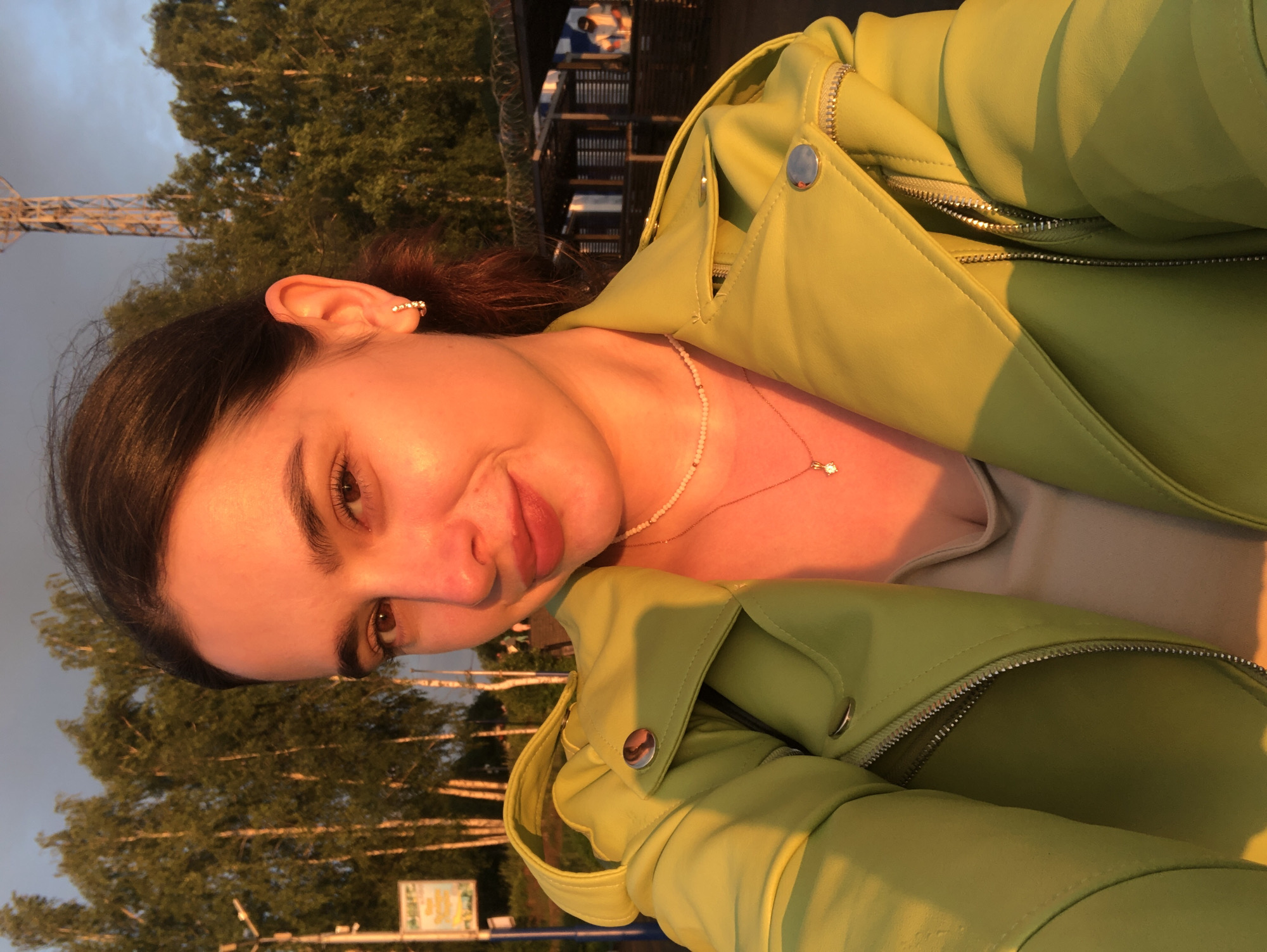 A young woman with dark hair, wearing a lime green leather jacket and a simple necklace, is captured in a selfie outdoors. The setting appears to be a park or recreational area in Chaikovsky, Russia, suggested by the presence of birch trees and what looks like a tall radio tower in the background. The warm golden light of sunset illuminates the scene, casting a soft glow on the woman's face and clothes. In the blurred background, structures that resemble food stalls or small buildings are visible, along with a few other people in the distance. A sign with Cyrillic text is also present on the left side of the image.
