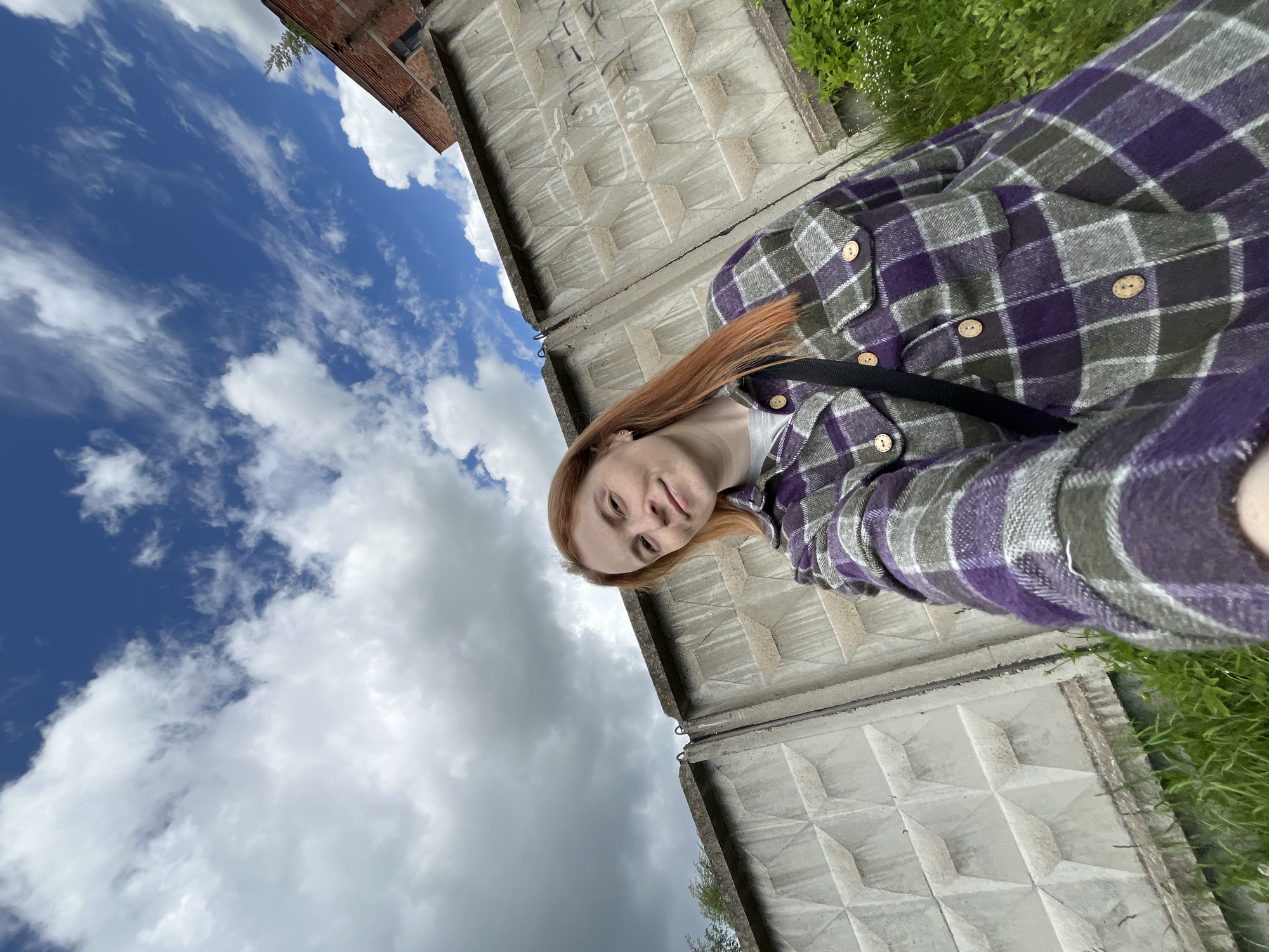 A woman with red hair, wearing a purple and gray plaid shirt with wooden buttons and a black strap over her shoulder, is taking a selfie outside in what appears to be Igra, Russia. She is positioned in front of a gray concrete wall with a geometric pattern. To the left of the wall, the sky is visible with a mix of blue patches and white clouds. In the upper left corner, there is a portion of a brick building. Some greenery is seen to the upper right of the wall.