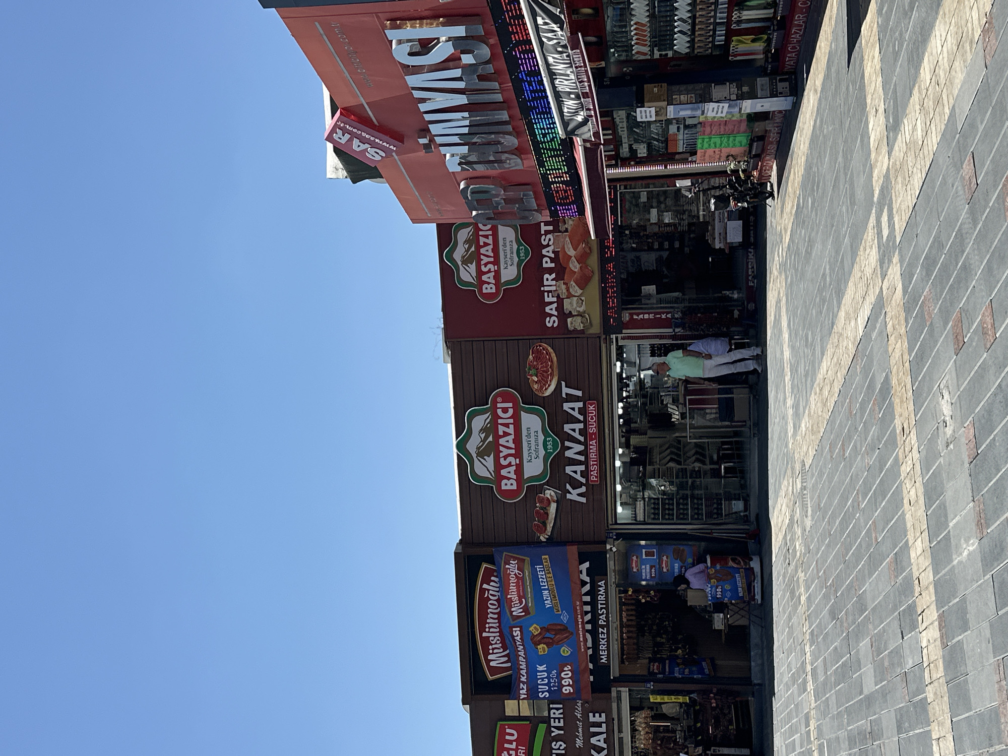 In the city of Kayseri, Turkey, a street scene unfolds under a clear blue sky. The foreground features a patterned pavement made of dark gray and light tan rectangular bricks. Beyond the pavement, a row of shops presents a vibrant facade of advertisements and signage.
One prominent shop displays a large banner for "Müslümoğlu" featuring images of cured meats and the text "YAZIN LEZZETİ MÜSLÜMOĞLU İLE BAŞLAR" (Summer's taste begins with Müslümoğlu). Adjacent to it, a red sign for "BAŞYAZICI" with an image of mountains reads "Kayseri'den Sofranıza 1953" (From Kayseri to your table 1953). Below this, a sign for "KANAAT" advertises "PASTIRMA - SUCUK" (cured meat - sausage).
Further to the right, a large, illuminated sign spells out "GÜNAYSI" in bold silver lettering, with smaller text above and below it. A colorful LED sign runs horizontally across the facade of this shop. Another shop on the far right displays a wide variety of items, including what appear to be phone accessories or electronics, arranged on shelves and in boxes.
Several individuals are visible. A person in a light green shirt and white pants stands near a shop entrance. Another person, dressed in a pink top and dark pants, is seen inside a shop, interacting with a display. A motorcycle is parked in front of one of the establishments. Inside the shops, various products, including strings of cured meats, are displayed. The overall impression is a bustling commercial street in Kayseri.