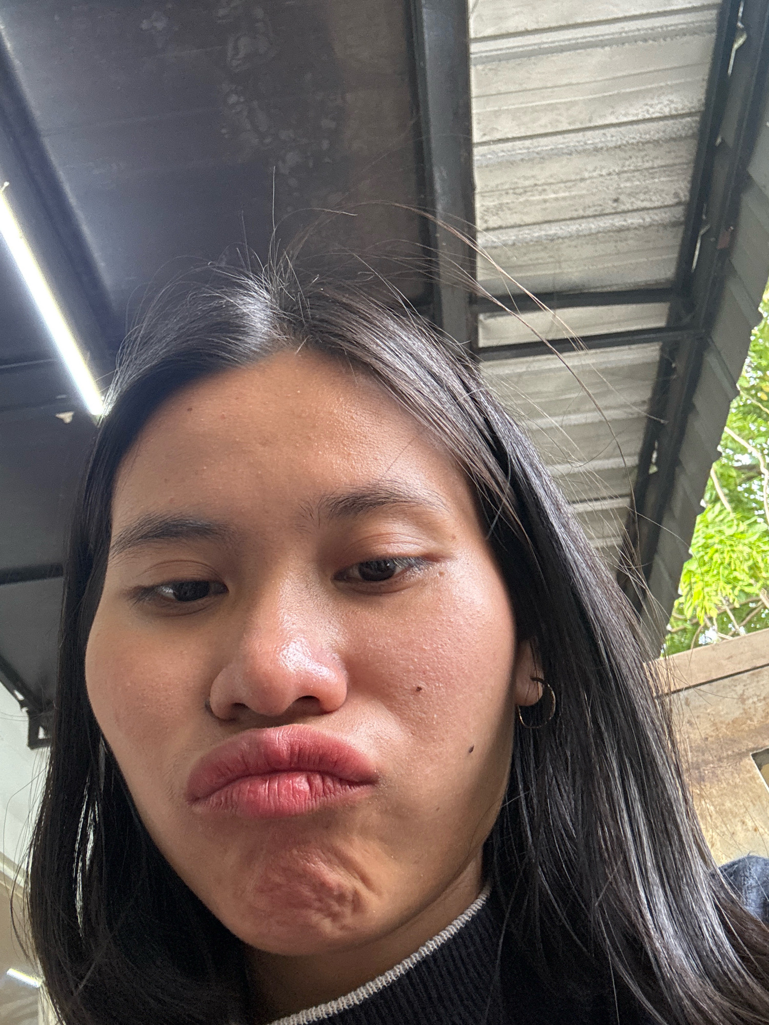 A close-up, low-angle selfie captures a young East Asian woman's face from the chin up. She has long, dark, straight hair that frames her face and extends down. Her skin appears fair with some blemishes. Her eyes are dark and looking slightly downwards. Her lips are pursed into a pout, and her chin is wrinkled. She is wearing a dark garment with a white trim at the neckline. A small hoop earring is visible in her left ear.
The background is an outdoor setting under a corrugated metal roof supported by dark metal beams. To the right of the woman, lush green foliage suggests trees or plants. The lighting suggests it is daytime. The scene appears to be located in East Jakarta, Indonesia, perhaps in a covered outdoor area.