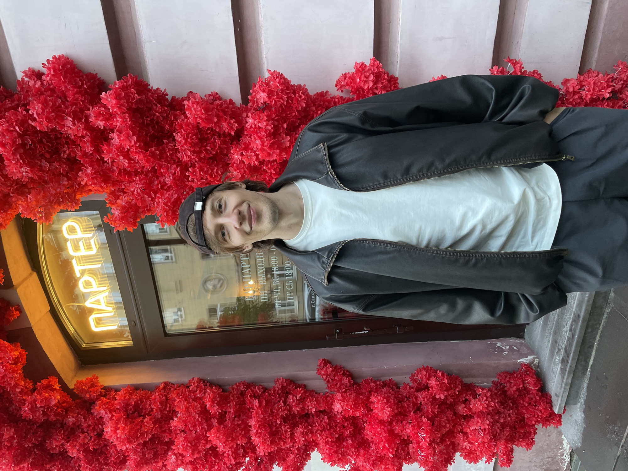 A full-body, eye-level shot shows a young man standing in front of a building in Saint Petersburg, Russia. He is wearing a dark bomber jacket over a white t-shirt and dark pants, with a baseball cap on his head. The building's entrance is framed by dense clusters of bright red decorative flowers. Above the entrance, a neon sign in Cyrillic reads "ПАРТЕР". The building has a glass door with text visible on it, including what appears to be business hours. The man is looking directly at the camera with a slight smile. The facade of the building is a pale pink color.