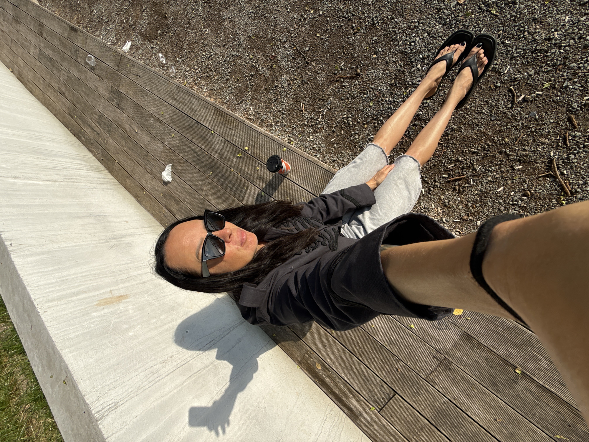A selfie is taken by a woman sitting on a wooden bench with her legs outstretched. She is wearing a black hooded jacket and grey shorts. She has long black hair and sunglasses. A coffee cup is placed on the bench beside her. The bench is situated next to a grey concrete wall and a gravel ground. The scene appears to be outdoors, possibly in a park or public space in Chaikovsky, Russia.