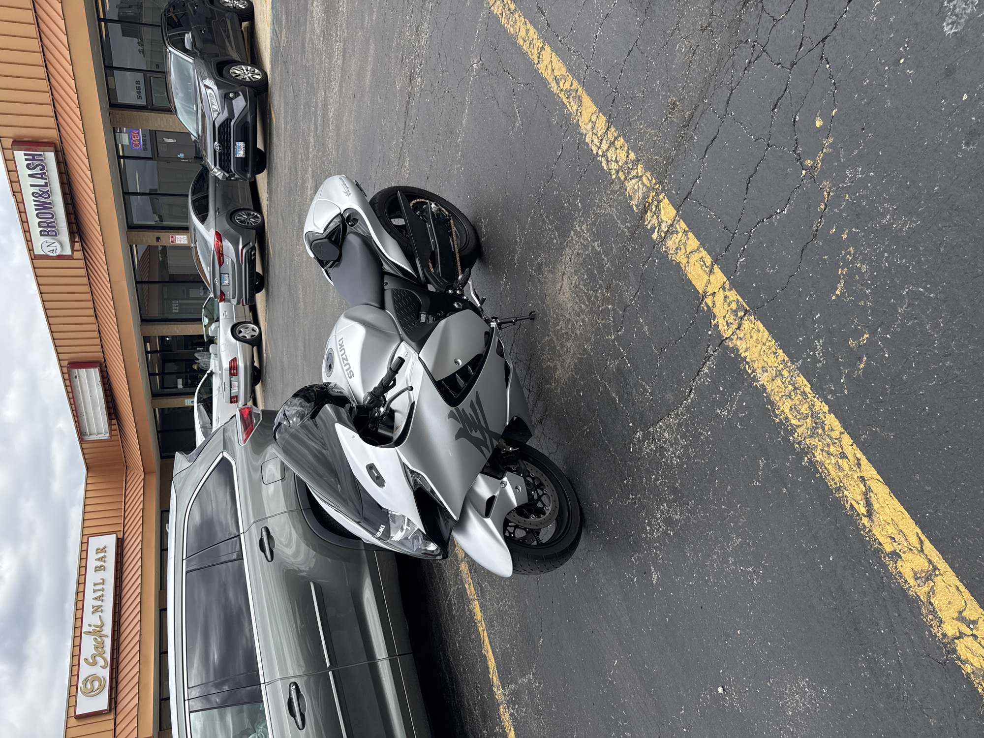 The image shows a silver Suzuki Hayabusa motorcycle parked on a black asphalt parking lot next to a gray minivan. The parking lot has yellow painted lines. In the background, a building houses businesses including "Sacki Nail Bar" and "AN Brow & Lash". Several cars are parked in the parking lot. The location of the scene is in the city of St. Charles, United States of America. The image suggests an urban or suburban area.