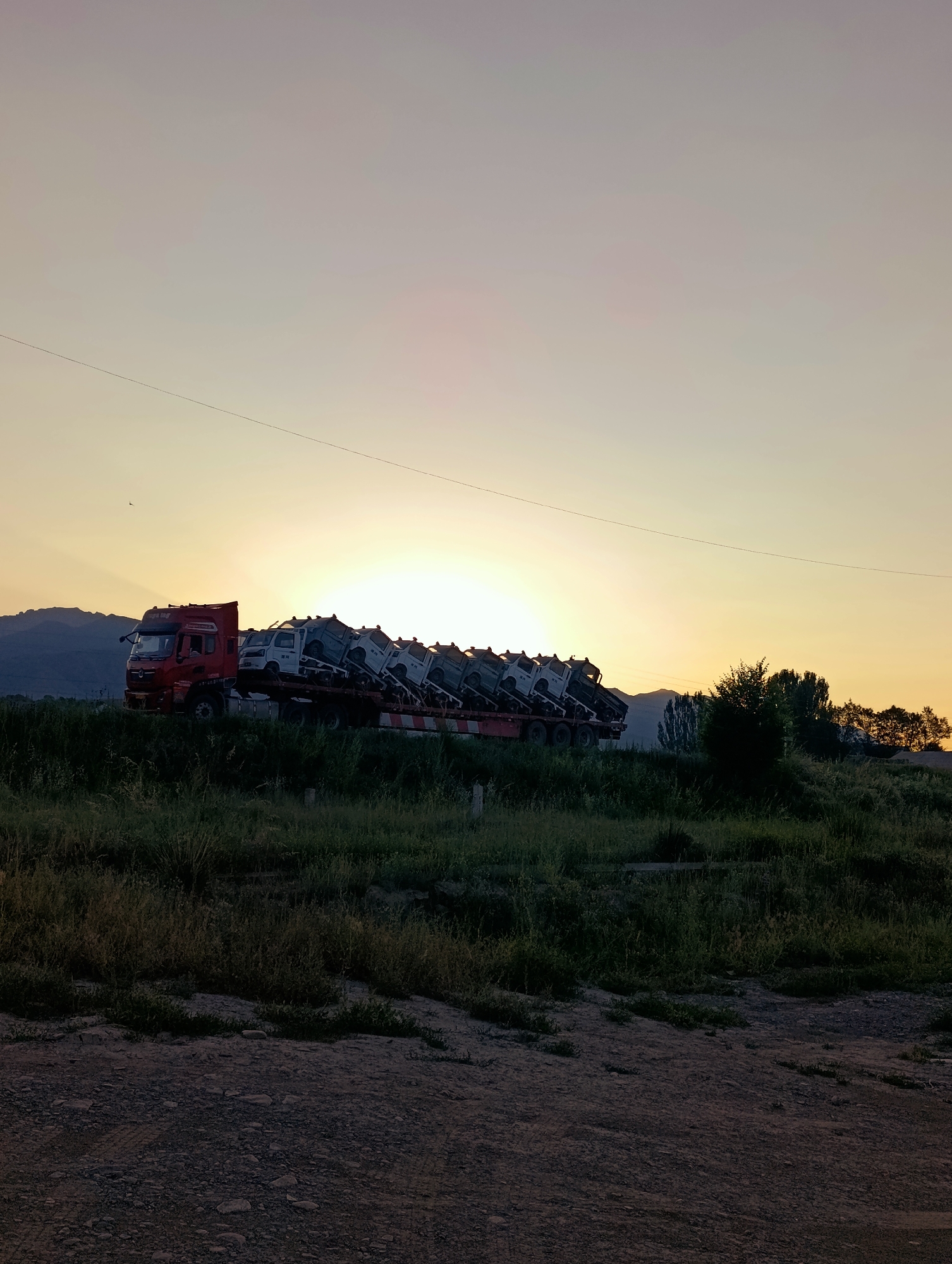 A wide shot captures a semi-trailer truck carrying several smaller trucks on its flatbed, silhouetted against a sunset sky. The truck is positioned on a road with a grassy embankment to its right and a dirt and gravel foreground. In the distance, mountains are visible against the horizon. The sky is a gradient of soft yellow, orange, and purple hues. A thin power line stretches diagonally across the upper portion of the frame. The scene is set in the city of Kyzyl-Jyldyz, Kyrgyzstan.