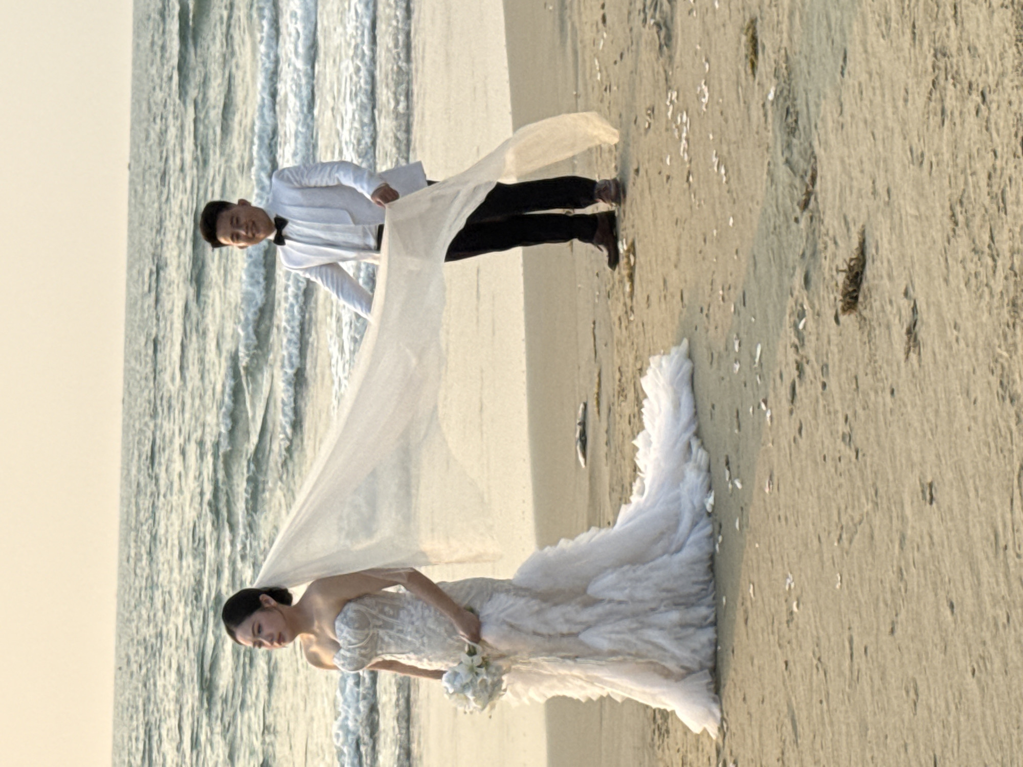 A wedding photo shows a couple on a beach in Đà Nẵng, Vietnam. The bride is wearing a white, strapless, mermaid-style wedding dress and holding a white bouquet. The groom is wearing a white tuxedo jacket, black bow tie, and black pants. They are standing on a sandy beach near the ocean. The groom is holding the bride's white veil, which is billowing in the wind. The beach is light tan, and the ocean is a light blue-gray. The sky is a pale yellow.