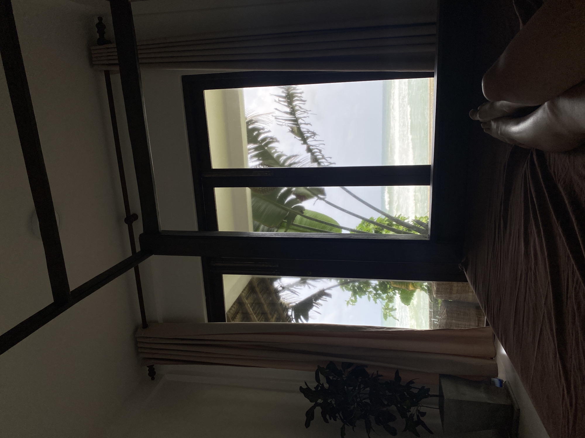 From a low-angle perspective inside a room, a dark wooden four-poster bed frame is visible. A window divided by a horizontal beam frames an ocean view with palm trees and foliage visible outside. The ocean appears light and the sky is overcast. Soft, light-colored drapes hang at the top and bottom of the window. A potted plant sits below the window. A person's legs and feet rest on a brown sheet on the bed. The location is Goviyapana, Sri Lanka.