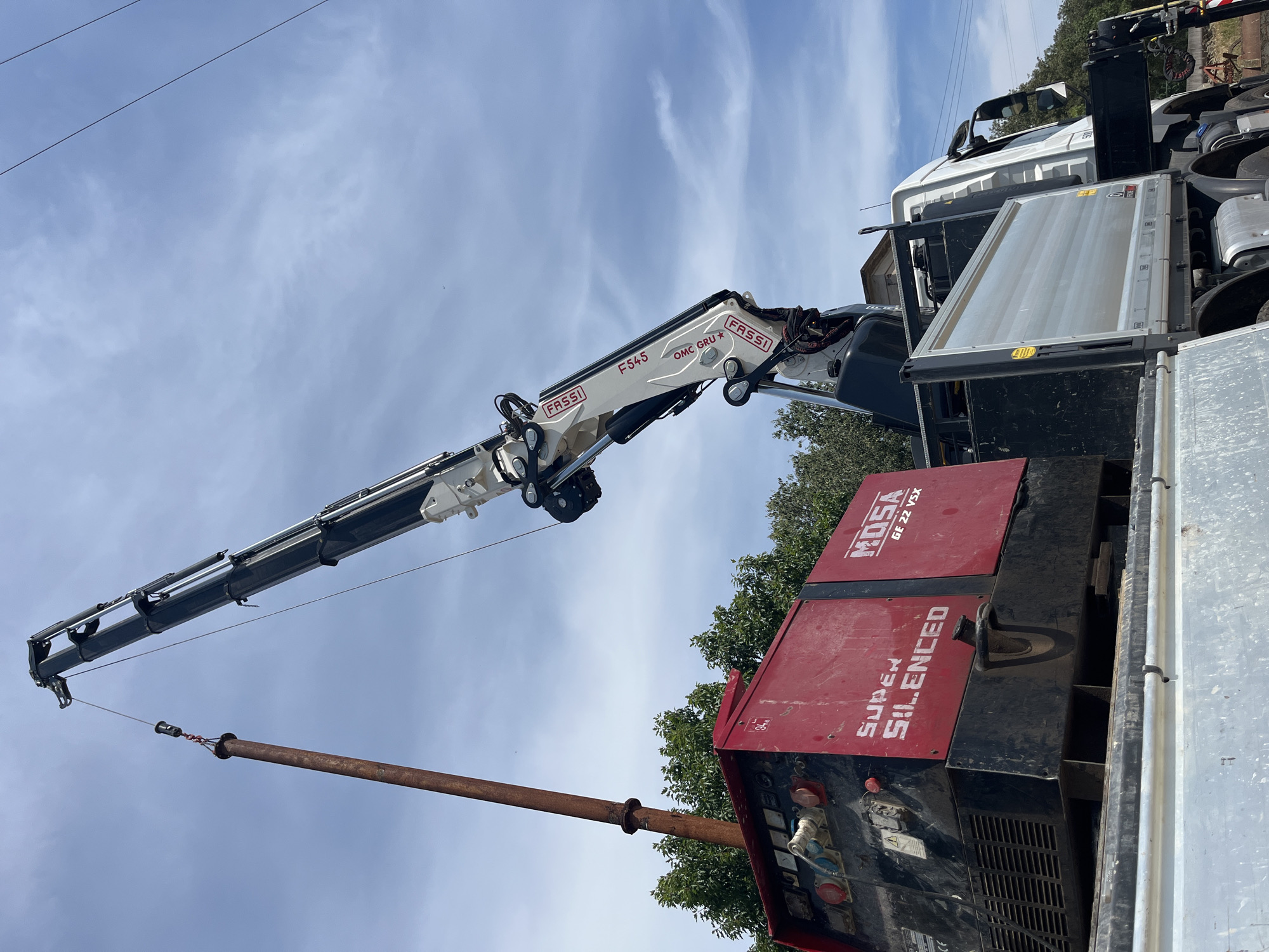 An exterior medium shot shows a large red and black industrial generator on the bed of a truck, positioned in the city of Castiglione delle Stiviere, Italy. A white crane is actively lifting a rusty metal pipe from above the generator. The pipe is suspended by a cable attached to a metal hook. The generator has "SUPER SILENCED" printed on its side in white letters and "MOSA GE 22 VSX" in red. The truck also has a silver cargo bed. The sky is blue with scattered white clouds, and green trees are visible in the background.