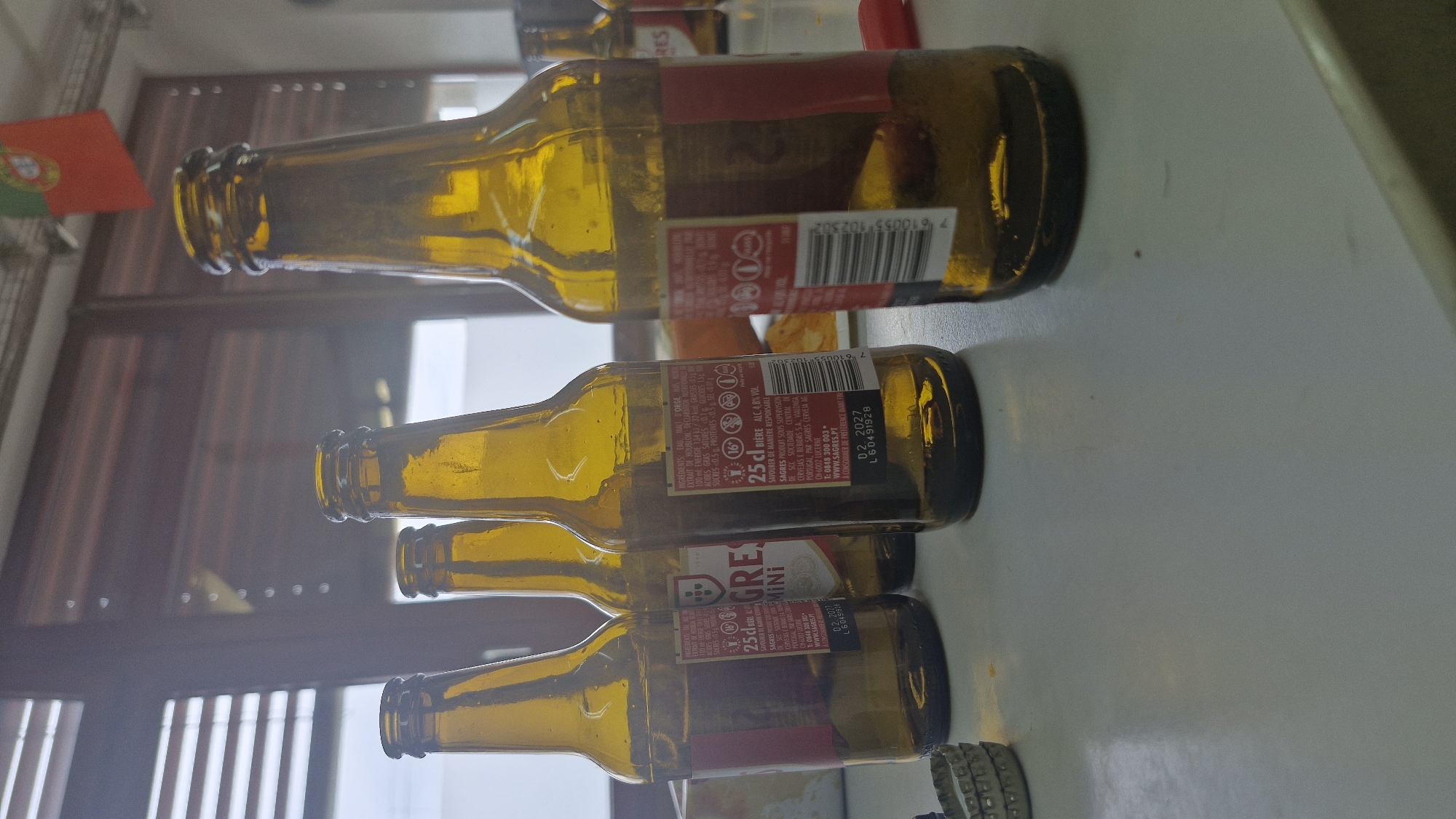 In Uetendorf, Switzerland, four amber glass beer bottles are arranged on a white, reflective surface. Three bottles are in the foreground, and one is slightly behind and to the right. The bottles are mostly empty, with some residual liquid visible at the bottom. Each bottle has a label with text and nutritional information. The bottle on the far left has a red label with white text. The second bottle from the left has a white label with a crest and text. The third bottle from the left has a label with text that includes "25cl BIÈRE ALC. 4.8% VOL." and a barcode. The bottle on the far right also has a red label with white text and a barcode. A Portuguese flag is visible in the background, hanging from a cord. The background also shows a window with horizontal blinds and a partially obscured outdoor scene. Several metal bottle caps are stacked to the left of the bottles.