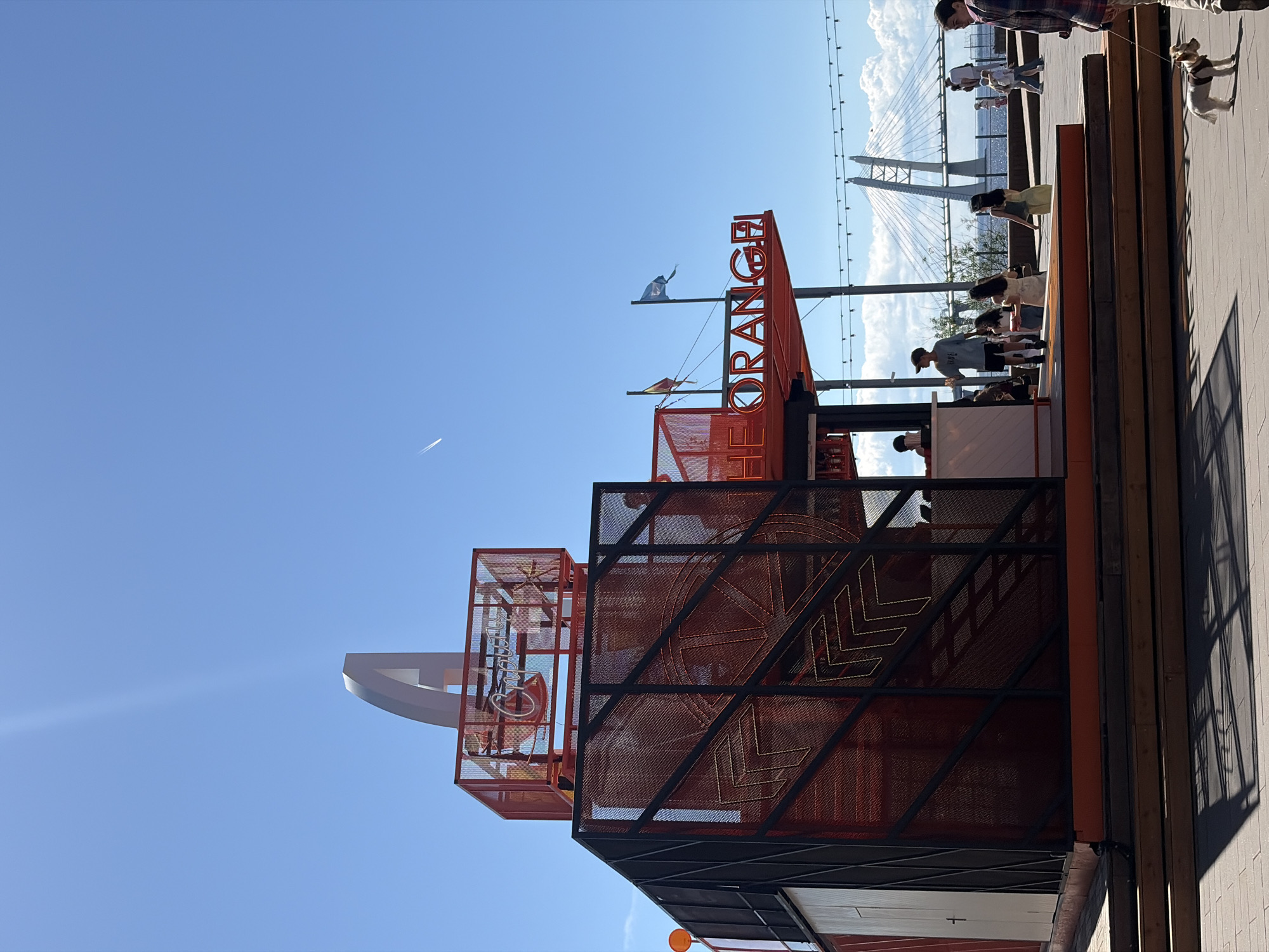 This image captures a bright, sunny outdoor scene in Saint Petersburg, Russia, featuring a multi-level structure identified as "THE ORANGE" against a backdrop of the Western High-Speed Diameter (WHSD) bridge.
The central subject is a modern, angular structure predominantly clad in orange mesh and black framing. Its lowest level features a white counter where staff members are visible behind it, with customers standing in front. Above this, a platform bears large, illuminated orange letters spelling "THE ORANGE." Two flags fly from this level: one with horizontal stripes of red, yellow, and red, and another blue and white flag. The top section of the structure includes an orange mesh box displaying a white neon sign that reads "Газпром" (Gazprom), topped by a prominent, stylized white or light-grey "A" or arch-like sculpture. Orange spherical lighting elements and yellow neon arrow patterns are integrated into the structure's design. The base of the structure is surrounded by several tiers of wooden decking, leading down to a paved ground surface.
Multiple people are present: a person in a dark cap and grey shirt stands near the counter. Several women in light-colored clothing are distributed around the structure and walking in the mid-ground. On the far right in the foreground, a person, partially cropped from the frame, walks a small, light-colored dog on a leash across the sun-drenched paving stones. The dog is light brown or cream-colored with short fur.
The background features a vast body of water, likely the Neva River or Gulf of Finland, under a clear, intense blue sky. The prominent white, cable-stayed bridge of the Western High-Speed Diameter is visible spanning the water. A line of white cumulus clouds is present near the horizon above the water. A faint airplane contrail streaks across the upper left portion of the sky. A string of decorative outdoor lights is suspended horizontally in the mid-ground, above the water. Harsh shadows cast by the structure and people on the paved ground indicate bright, direct sunlight.