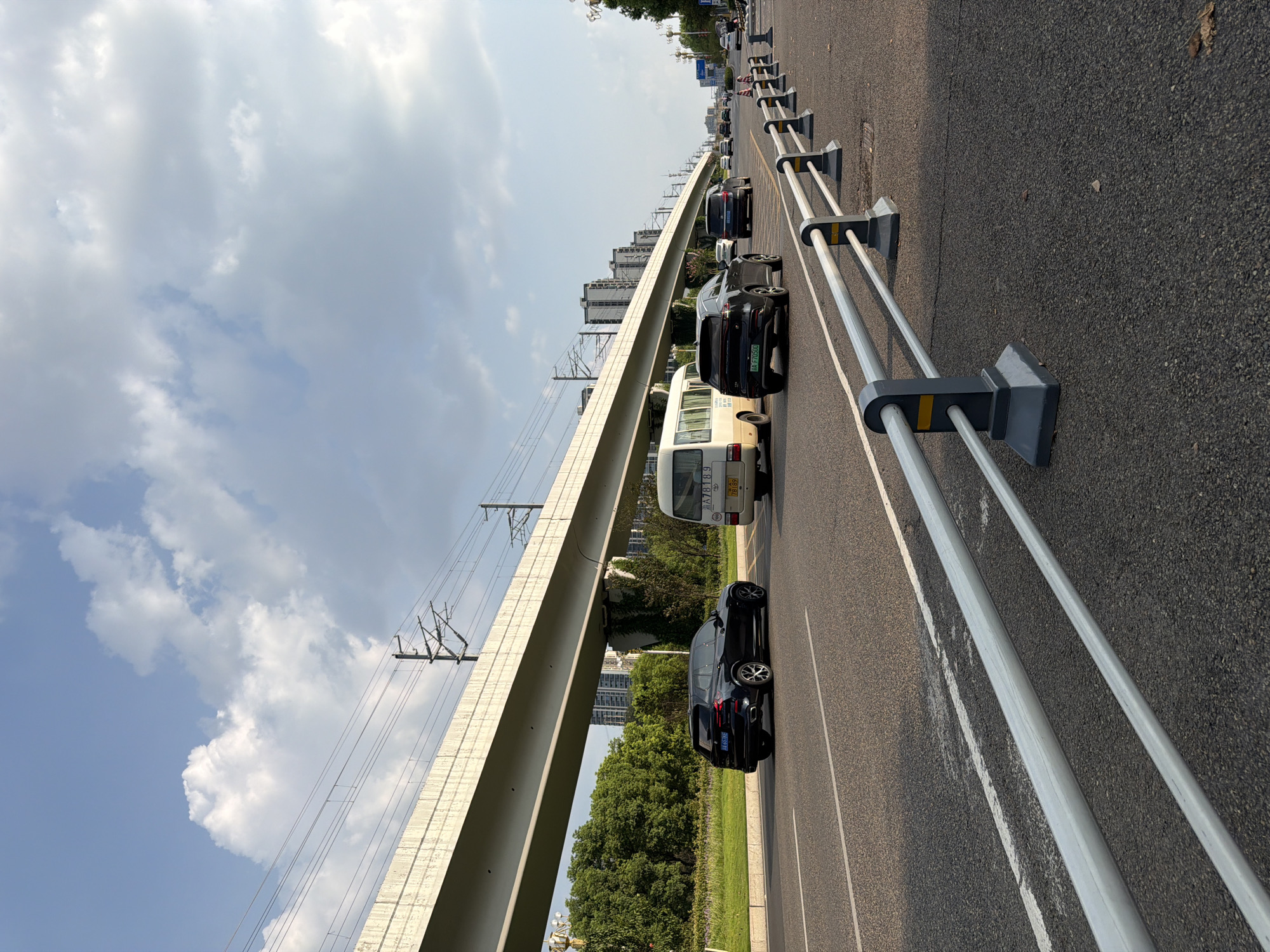 The image depicts a street scene in Jiangjin, China, likely during daylight hours given the clear sky with scattered clouds and the presence of shadows. An elevated roadway or railway track supported by concrete pillars runs diagonally across the frame from left to right, with overhead power lines visible. Below the elevated structure, a multi-lane road is in motion. Several vehicles are present: a dark-colored SUV, a white minivan, and other cars further down the road. The road is lined with a barrier system consisting of grey posts with white and yellow accents. Green trees and residential or office buildings are visible in the background, suggesting an urban or suburban environment. The overall impression is one of a functional, modern city infrastructure with active traffic.
