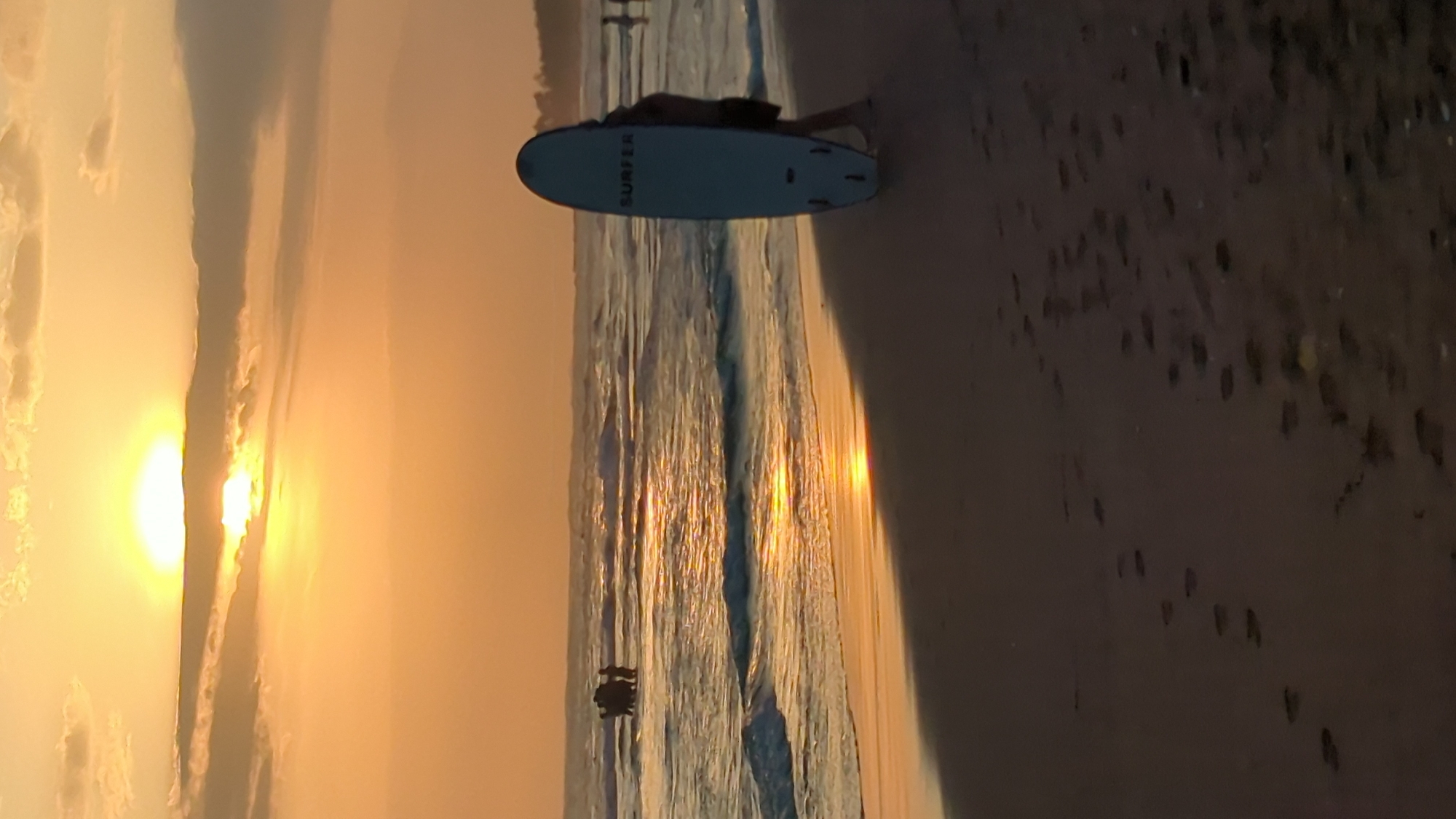 The image captures a serene beach scene at sunset in North Kuta, Indonesia. The golden light of the setting sun bathes the sky in warm hues of orange and yellow, casting a radiant glow across the water and the sandy shore. Wispy clouds are scattered across the sky, adding texture to the sunset.
In the foreground, a light blue surfboard lies on the sand, with the word "SURFER" visible in black lettering. A person is lying on the surfboard, partially visible, suggesting they are either resting or preparing to surf. Further out, a small dark object, possibly a boat or a person swimming, is visible in the shallow water. The wet sand near the waterline reflects the sun's vibrant colors, creating shimmering patterns. Footprints are scattered across the dry sand, hinting at the presence of other beachgoers. The gentle waves of the ocean roll onto the shore, creating a peaceful and tranquil atmosphere. The overall mood is one of calm and relaxation, typical of a beach at dusk.