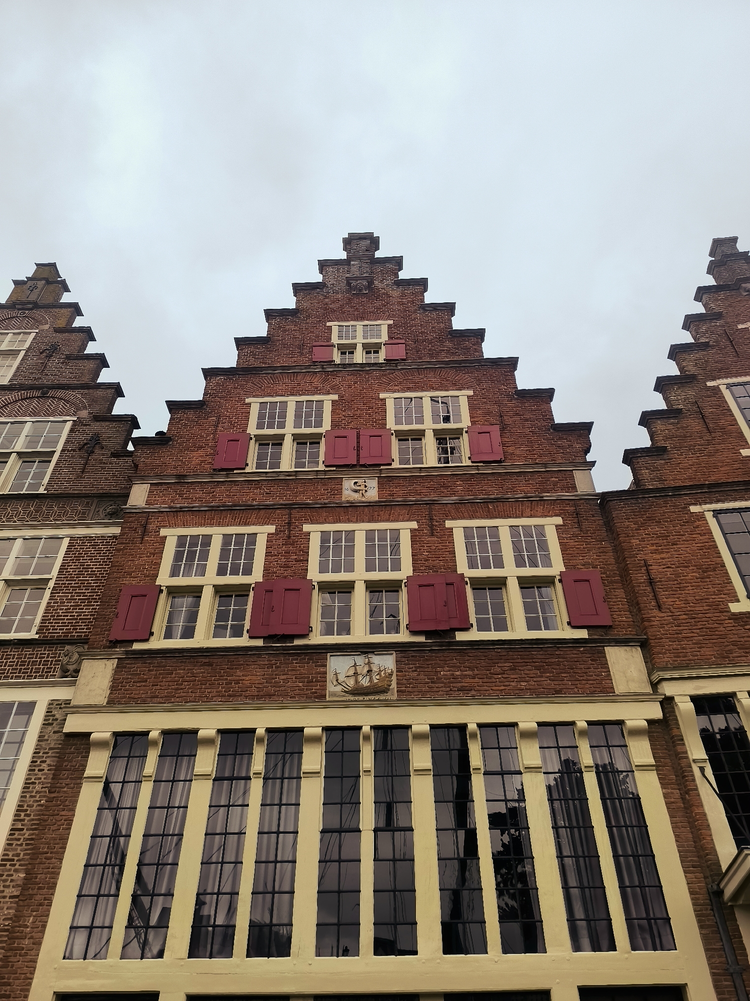 This is a low-angle, exterior, daytime, full shot of several Dutch stepped gable buildings in Hoorn, Netherlands. The buildings are constructed of red brick with white trim and a yellow band above the lower windows. Each building features multiple stories with numerous windows, many adorned with red shutters. At the top of the central building, a decorative crest and the inscription "1677" are visible. Above the lower windows of the central building, a small relief depicting a sailing ship is mounted. The sky is overcast and gray.