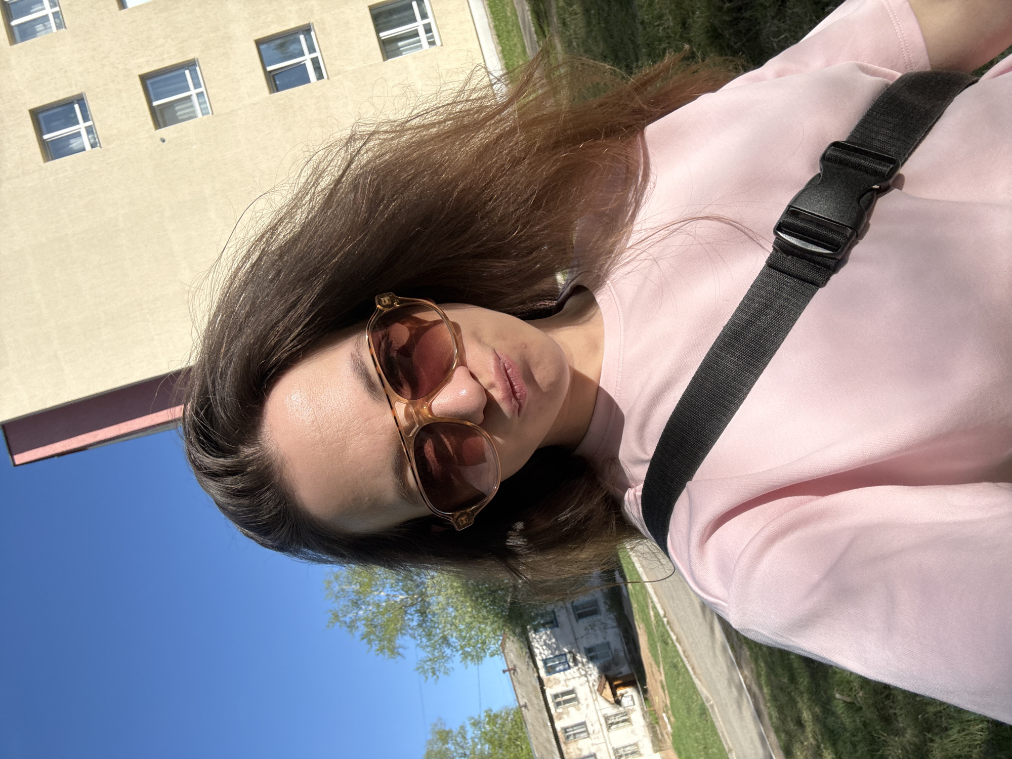 The image shows a woman with brown hair and sunglasses, wearing a light pink top with a black strap across her chest. She is outdoors on a sunny day in what appears to be an urban environment. In the background, there are multi-story buildings with visible windows. Further in the distance, trees and greenery can be seen, indicating a park or a grassy area. The location is Chaikovsky, Russia.