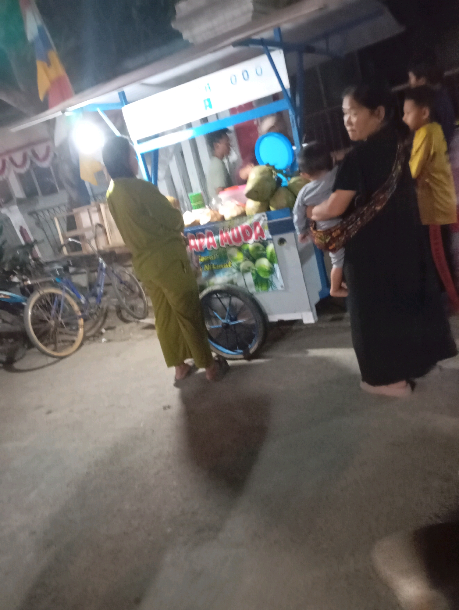 The image depicts a nighttime scene at a street food stall in Kecamatan Haurgeulis, Indonesia. A person in a green outfit stands in the foreground, facing away from the camera and towards the stall. Behind them, a woman in a black dress carries a baby, with a child in a yellow shirt standing to her right. The stall itself features a cart with a blue frame, displaying coconuts and possibly other produce. The ground is paved and appears to be a sidewalk or street. Several bicycles are visible in the background, suggesting a common mode of transportation in the area. The lighting suggests it is evening or nighttime, with the food stall illuminated by artificial lights.