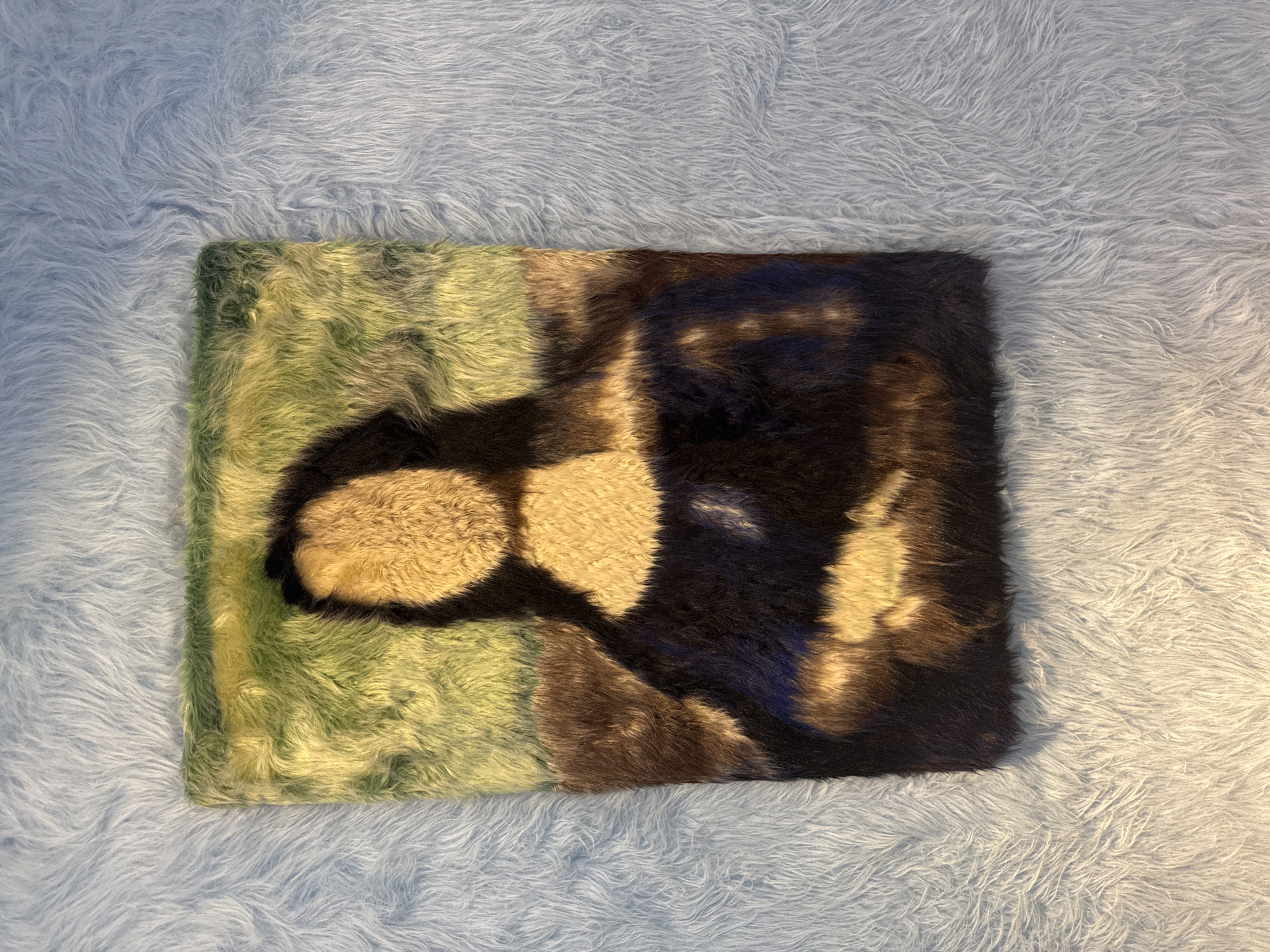 The image shows a fur rendition of the Mona Lisa placed on a light blue, fluffy surface. The fur artwork is rectangular and features the familiar portrait with its characteristic smile. The background of the artwork is depicted in various shades of green and brown, while the subject's face and hands are rendered in lighter, more yellowish fur. The Mona Lisa artwork is surrounded by a textured, light blue, furry material. The lighting casts a shadow on the bottom left portion of the blue surface, indicating a light source from the top left. There is no information in the image that can be used to determine its location in Munich, Germany.