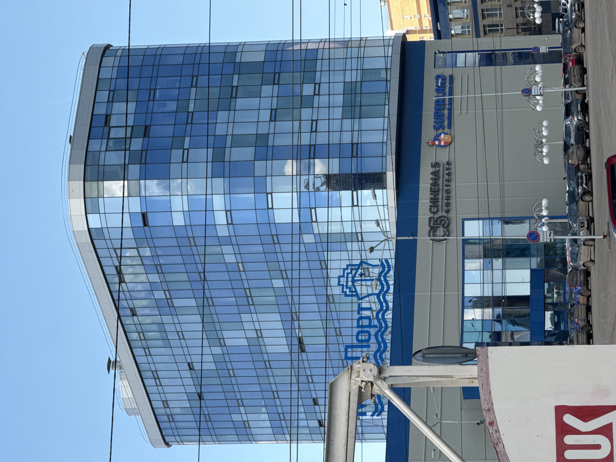 The image shows a tall, modern building with a curved, glass facade reflecting the sky. The building appears to be a cinema, identifiable by the signage "С5 СИНЕМА5 КИНОТЕАТР" and "SUPER C5" in Cyrillic script. A blue and white logo resembling a ship or water park is also visible. The building has blue trim and is located on a city street, with parked cars and streetlights alongside. Overhead, power lines are visible against the clear blue sky. Based on the signage, the building is located in Izhevsk, Russia.