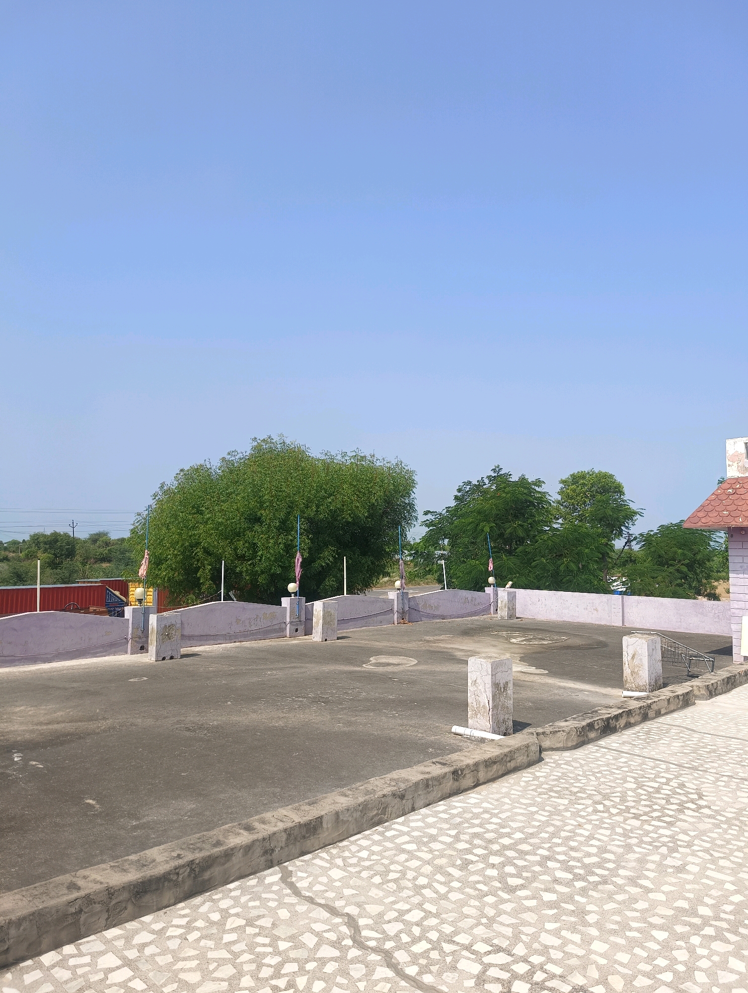 The image shows a rooftop view in Chhapri Bari, India. The foreground features a textured tiled floor with a concrete border. Behind the border is a darker concrete surface, likely the main rooftop area. A low, light-colored wall with occasional square concrete posts lines the edge of this area. Two of these posts are visible, one with a pipe extending from its base. The wall has some flags with pink cloth attached to poles on either side of it. In the middle ground, several lush green trees are prominent, with a larger, bushier tree dominating the center. Beyond the trees, a red shipping container is visible, along with utility poles and more vegetation. The sky is a clear, bright blue, indicating a sunny day. A portion of a building with a red tiled roof is visible on the far right side of the image. There are no people visible in the scene.