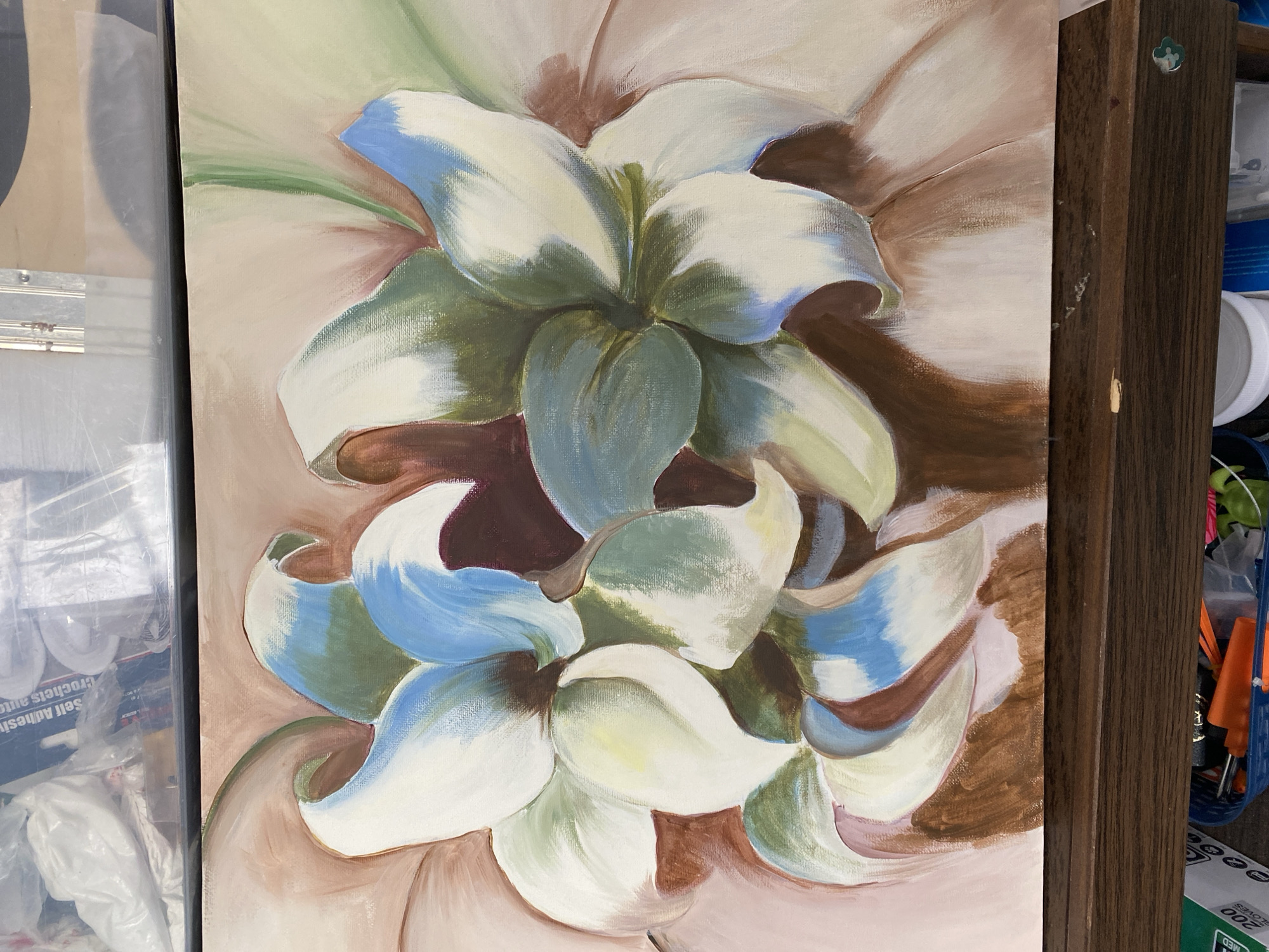 The image is a photograph of a painting of lilies on a canvas, displayed on a wooden easel. The painting features large, stylized lily blossoms in white, green, and blue hues, set against a pale pink and brown background. The brushstrokes are visible, suggesting an impressionistic style. The top portion of the image shows a blurry background of plastic-wrapped items and some shelving. The bottom portion shows the wooden frame of the easel and a glimpse of a blue basket containing tools and containers. The scene appears to be an indoor setting, possibly an art studio or a room where the painting is being worked on or displayed. There is no indication of the city of Miami, United States, within the image or its context.