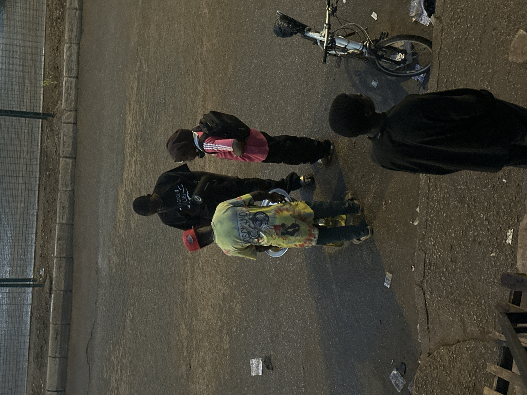 Four young males are gathered on a paved street in Bukuru, Nigeria. One male, wearing a black t-shirt with white text that reads "GOOD LIFE," stands facing right. Another male, to his left, wears a red baseball cap, a tie-dye t-shirt with a cartoon character and text, and dark pants, holding a silver bowl. To the right of the first male, a third individual wears a pink t-shirt with white stripes, black pants, and a backpack, facing left. A fourth male, at the bottom right of the image, faces away from the camera wearing a black sweatshirt. A white bicycle with a black seat and handlebars is parked near the right edge of the frame. The ground is littered with debris, including plastic wrappers and paper. The background features a chain-link fence and a curb. The lighting suggests it is either early morning or late evening.