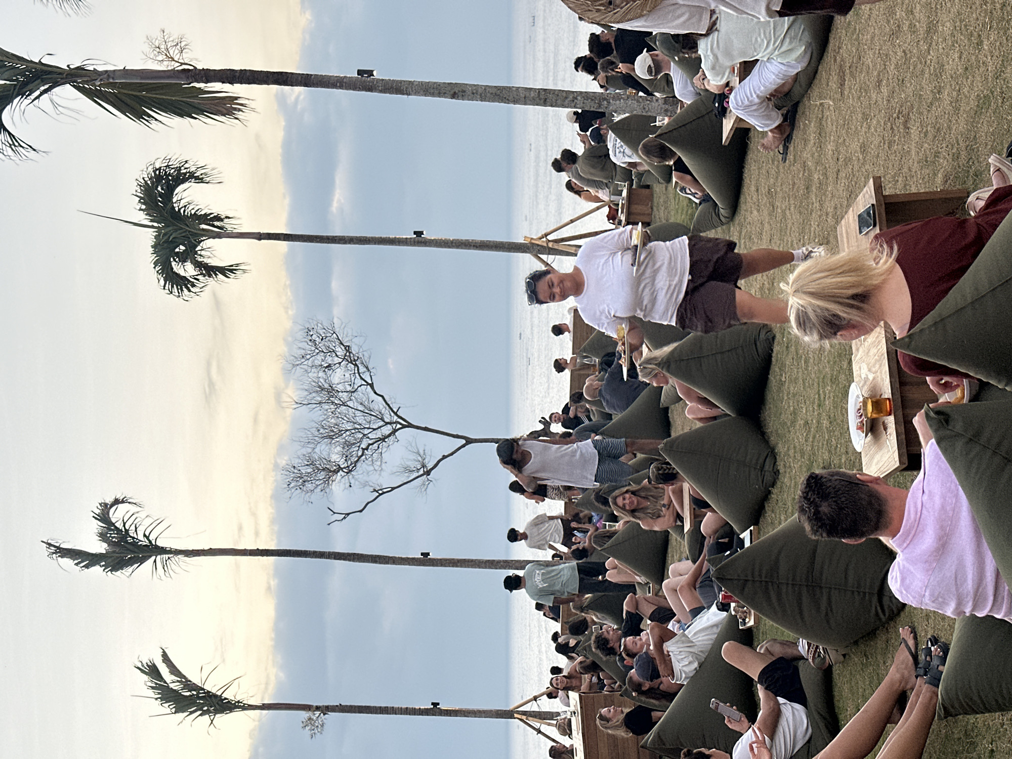 The image depicts an outdoor gathering at a beachside venue in Kuta Selatan, Indonesia, likely during the late afternoon or early evening, given the soft lighting and cloudy sky. Numerous people are seated on large, olive-green beanbag chairs arranged on a grassy slope leading towards the ocean. In the foreground, a woman wearing a white t-shirt and brown shorts is carrying two plates of food, seemingly serving patrons. To her right, a man in a light pink button-down shirt is seated, facing away from the camera, with a small table holding a drink and a plate of food in front of him. Further back, more individuals are scattered across the beanbags, some interacting with each other, others looking towards the sea. Several tall palm trees are visible, framing the scene against the backdrop of the ocean and the sky. The general atmosphere appears relaxed and social, suggesting a restaurant or bar setting.