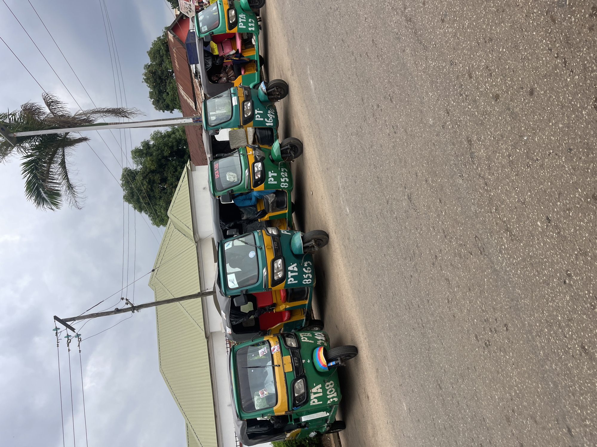 The image depicts a street scene in Jos, Nigeria, featuring a line of five green and yellow auto-rickshaws, commonly known as "Keke Napep" or "Marwa," parked on the side of a paved road. The auto-rickshaws are identified with numbers and markings such as "PTA 3108," "PTA 8565," "PT-16470," and "PTA 115." Several drivers and passengers are visible inside and around the vehicles. The background shows a building with a light green, corrugated metal roof and a white facade, along with trees and overhead power lines supported by a wooden pole. The sky is overcast with gray clouds.