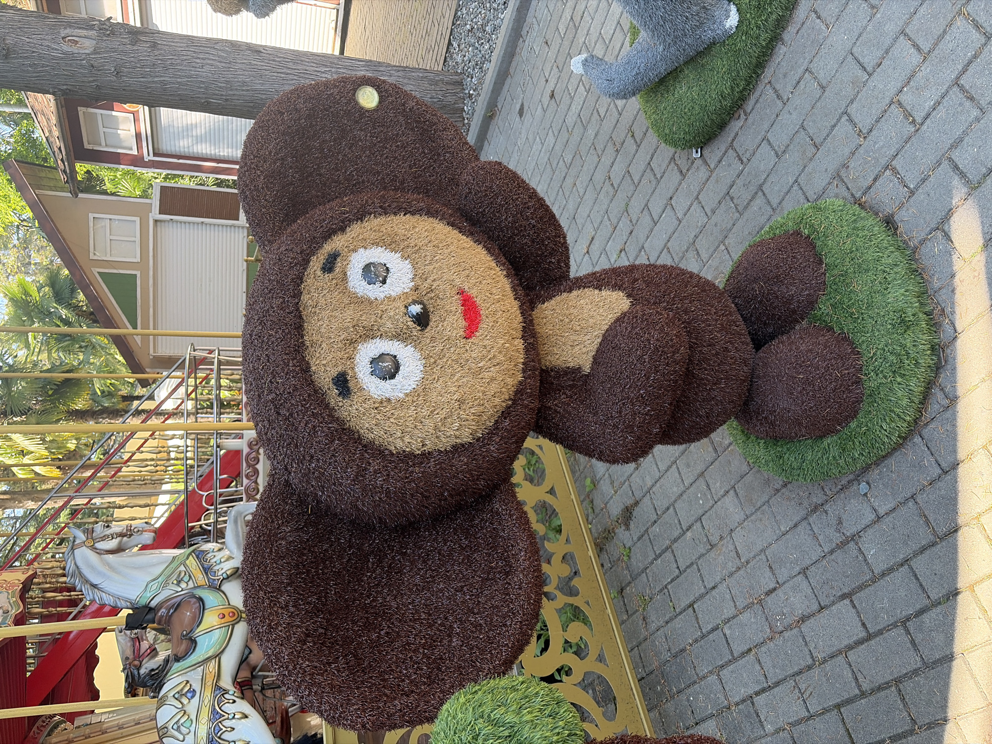 A Cheburashka topiary is displayed in a park in Sochi, Russia, in front of a carousel. The Cheburashka figure is made of brown and tan textured material, with large brown ears, white eyes with black pupils, and a red mouth. It stands on a green, artificial turf base. Behind the Cheburashka, a portion of a carousel is visible, featuring a white horse with a brown saddle and colorful decorations. The carousel appears to be made of wood and metal. The ground is paved with grey interlocking stones, and scattered pine needles are visible. In the background, there are trees and buildings with red and white facades and white roll-up doors. To the right of the Cheburashka, a grey animal topiary with its paw raised is partially visible on a similar green base. Sunlight casts shadows on the paving stones.