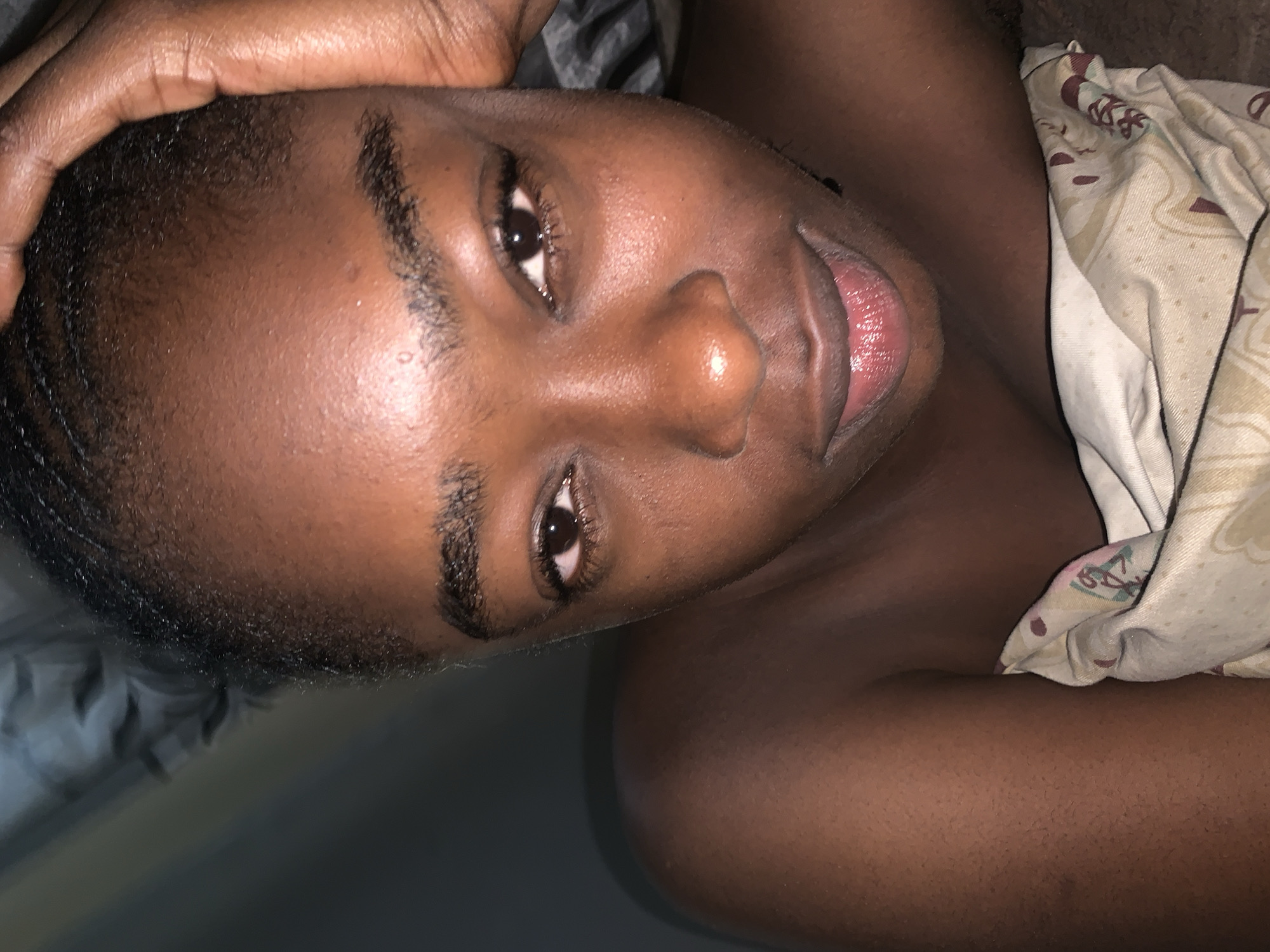 An intimate, close-up portrait shows a person with a dark skin tone lying down in a relaxed, reclined position. The individual's head and upper torso are oriented diagonally across the frame, with their gaze directed towards the viewer. They have short, dark hair, natural dark eyebrows, and dark eyes. Their lips are parted in a gentle, almost shy smile, revealing the tip of their tongue. One hand, also with dark skin, rests above their forehead or on the crown of their head.
The setting appears to be indoors, likely a bedroom or resting area, with subdued lighting that suggests either evening, early morning, or a dimly lit room in Rafin Par, Nigeria. A light-colored blanket or sheet with a repeating pattern, featuring muted floral or abstract motifs in colors such as maroon and beige, covers the person's right side. Another solid-colored fabric, possibly a grey or dark blue sheet or pillow, is visible in the background on the left. The person's skin exhibits a natural, subtle sheen. The overall impression is one of calm and repose, captured in a personal moment. No text is visible in the image.
