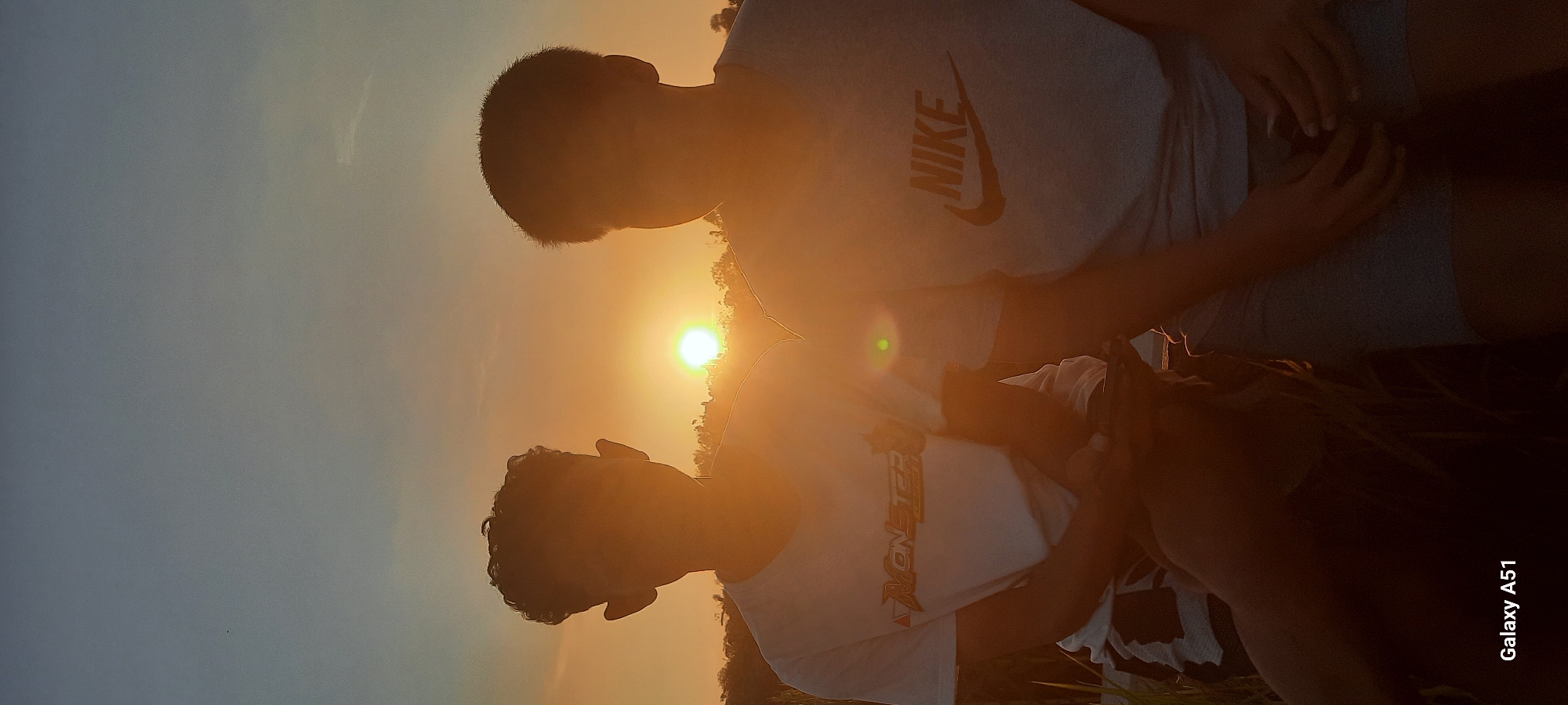 Two individuals are captured in a waist-up portrait, silhouetted against a setting sun. The person on the left wears a white t-shirt with "MONSTER" text, while the person on the right sports a grey t-shirt with a "NIKE" logo. Both individuals are seated and appear to be holding something in their hands, though the objects are not clearly discernible due to the low light conditions and overexposure from the sun. The background suggests an outdoor environment, possibly a field, with indistinct foliage and trees visible in the distance. The overall lighting is warm and golden due to the sunset. The image is tagged with "Galaxy A51," indicating the device used for capture. The location is identified as Duren, Indonesia.